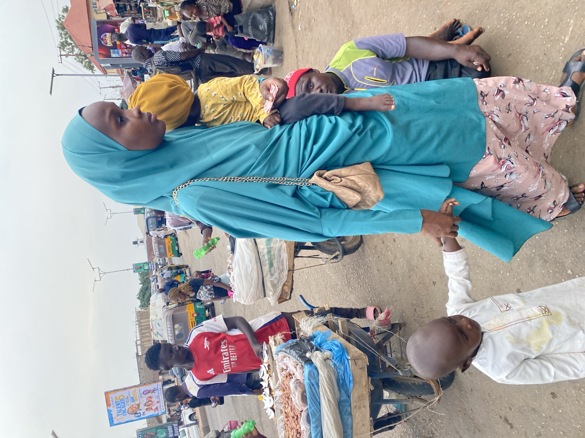 The image depicts a street scene in Gura Top, Nigeria. A woman in a blue hijab is seated, with children lying on her lap and next to her. A man in an Arsenal jersey stands beside a cart laden with goods. Another man in a white robe is lying on the ground with his head near the cart. Additional people and vehicles are visible in the background. Street signs, including one advertising "Talents Uncaged," are also present.