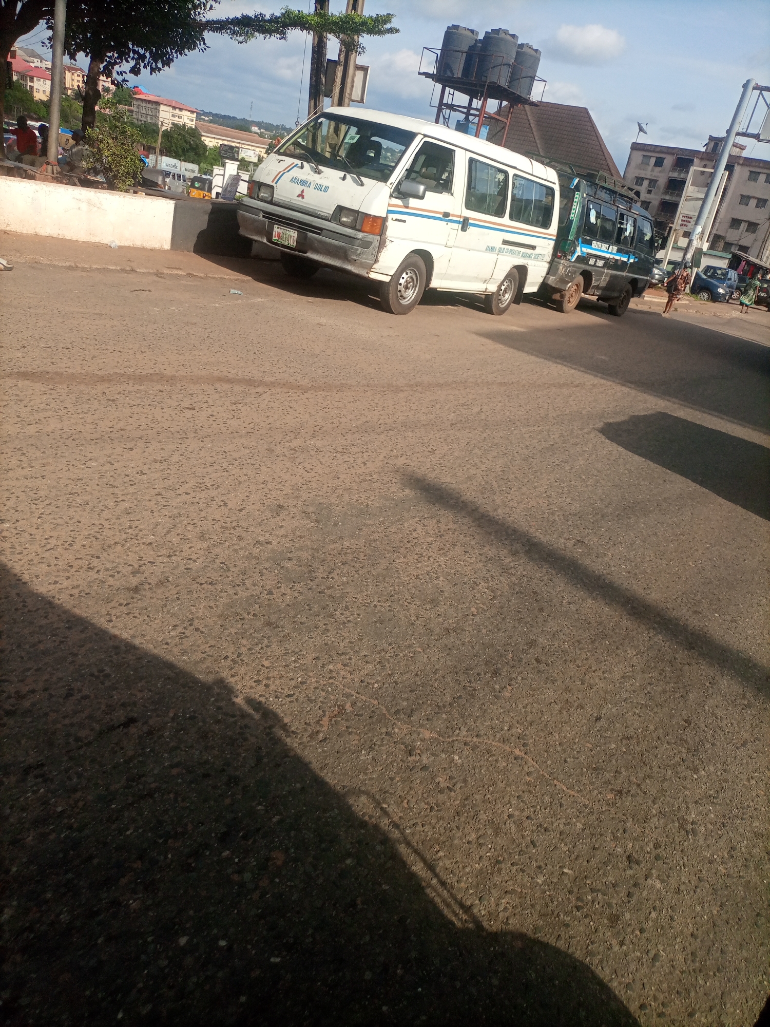 The image depicts a street scene in Nkpor, Nigeria, likely during the daytime under clear skies. The foreground is dominated by a rough asphalt road, with a large shadow cast from the left, suggesting late afternoon sun.
The central focus is on two vehicles parked on the road. The vehicle on the left is a white commercial minibus with "ANAMBRA SOLID" written in blue lettering on its side, and "AJARA AUTO" also visible. It has a license plate that appears to read "ANW 511 AA". To its right is a darker colored commercial vehicle, possibly a truck or another minibus, with writing on its side that is partially obscured but includes "BUS TRANSIT".
In the background, a white wall separates the road from a raised area where several people are seated or standing. Behind them, buildings of various heights and colors are visible, some appearing to be multi-story structures. Trees with lush green foliage are also present, suggesting an urban or semi-urban environment. Above the vehicles, a structure with several large black water tanks is mounted on a platform. The overall impression is a busy, lived-in street environment typical of a Nigerian town.