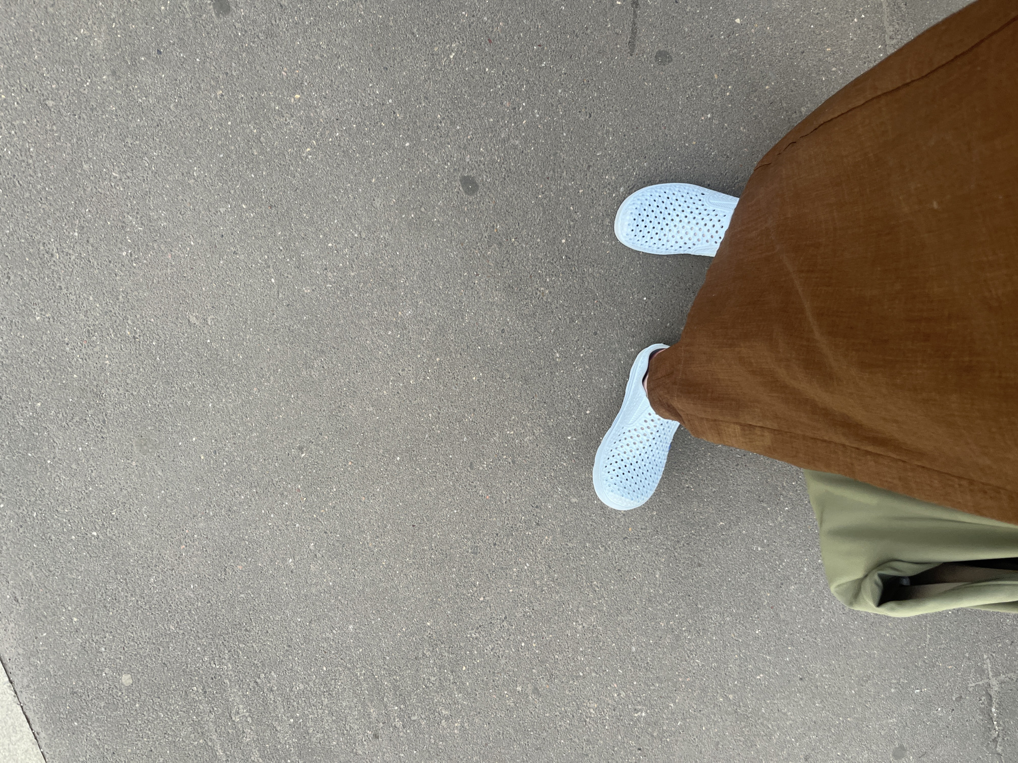 The image shows a downward-angled perspective of a person standing on a gray asphalt surface. The person is wearing brown trousers and light blue perforated shoes. The hem of what appears to be a green bag or garment is also visible near the bottom right of the image. The setting is a paved area, possibly a sidewalk or street. The location is not discernible from the image.