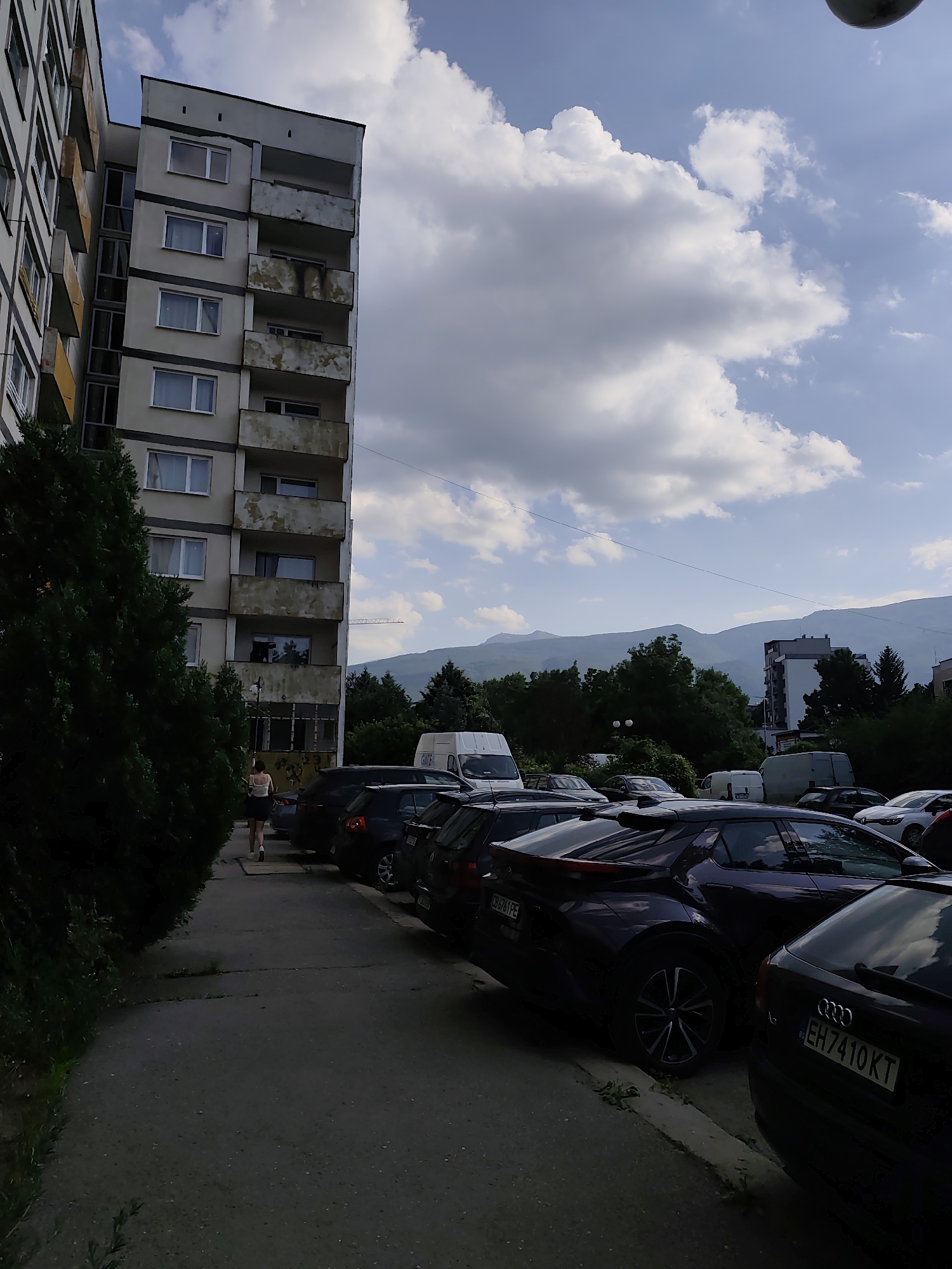 A vertical shot shows a parking lot in front of a Soviet-era apartment building. The building is tall and slender, with rows of balconies showing signs of age. The balconies are made of concrete and have yellowing railings. The building's facade is painted a pale gray. Adjacent to the building is a dense line of dark green trees.
A narrow sidewalk runs along the side of the building, and several cars are parked along the street. The cars are of various makes and models, and some are dark-colored. One car, a dark purple hatchback, is prominently featured in the foreground. It has a sporty design with a distinctive rear spoiler. The license plate is visible and reads "CB7871PE." Another car, a black Audi, is parked to the right, with its license plate visible as "EH7410KT." A woman in a white tank top and dark skirt is walking away from the camera on the sidewalk, her back to the viewer.
In the background, beyond the trees and cars, are low mountains under a partly cloudy sky. The clouds are large and white, with some darker, more dramatic formations. The sky is a clear blue, suggesting it is daytime, likely late afternoon or early evening due to the lighting conditions and the quality of the light on the mountains. The overall scene depicts a common urban environment, possibly in a residential area of Sofia, Bulgaria, given the architectural style and mountain backdrop. There is no strong indication of emotion, but the scene appears calm and ordinary.