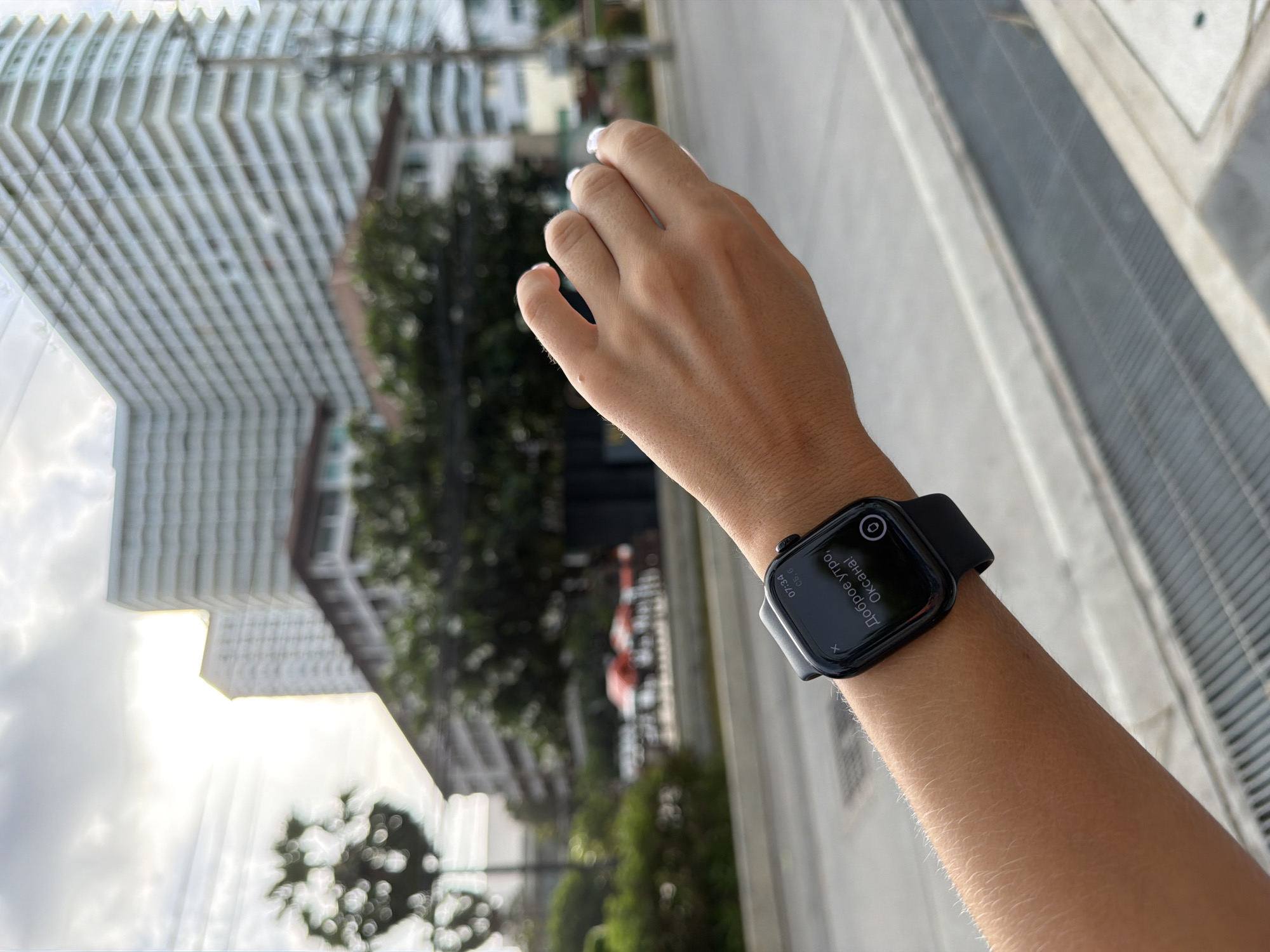 A close-up, vertical shot captures a light-skinned arm and hand wearing a black Apple Watch, set against a significantly blurred urban background. The arm extends from the lower-left, with the smartwatch positioned on the wrist. The watch face, illuminated, displays "07:34" and "СБ, 6" at the top right, with a notification below reading "Доброе утро, Оксана!" (Good morning, Oksana!). A small 'x' icon is visible at the top-left of the notification, and a circular icon appears at the bottom-right. The fingers of the hand are slightly curled, revealing white nail polish. Below the arm, a dark grey metal grate or drain runs horizontally, indicating a sidewalk or roadside. The background features various structures and natural elements rendered in a soft-focus bokeh. Dominating the upper right is a tall, light-colored multi-story building with numerous balconies or horizontal facade elements. Other buildings, including a lower-rise structure with a reddish-brown roof, are visible amidst dense green tropical foliage. The sky is bright and overcast, with a strong sun flare emanating from the upper-left center, suggesting a bright morning. Thin utility wires traverse the upper sky. The architectural style and tropical vegetation are consistent with an urban environment in Pattaya City, Thailand.