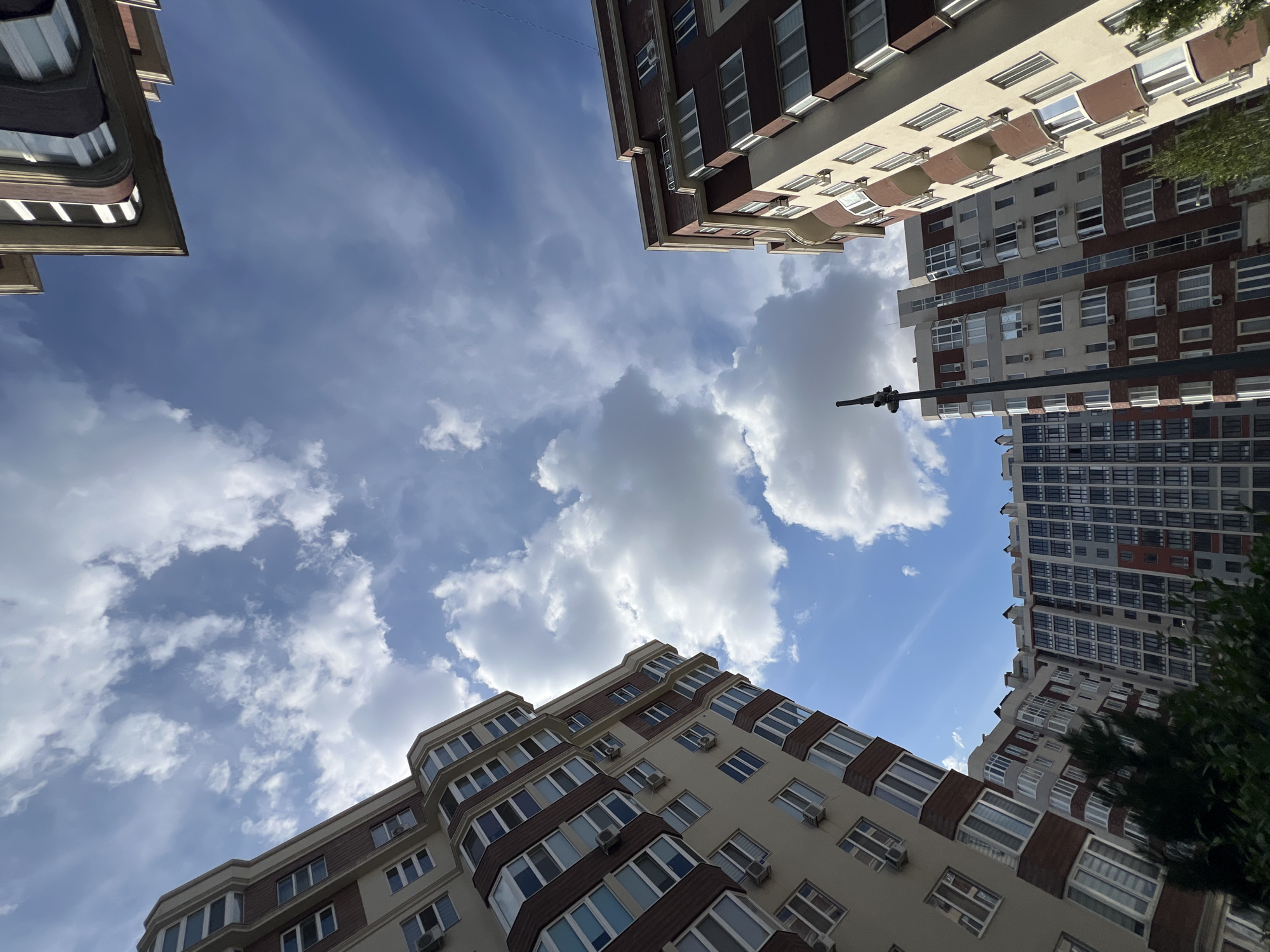This is a low-angle, vertical, outdoor, daylight shot of multiple multi-story apartment buildings in Kryzhanivka, Ukraine. The buildings are primarily light beige and dark red with numerous windows and balconies, some featuring air conditioning units. The sky above is blue with scattered white cumulus clouds. The buildings are angled and dominate the frame, with a street light visible in the middle ground. Some greenery from trees is present at the bottom of the frame. No people are visible in the image.