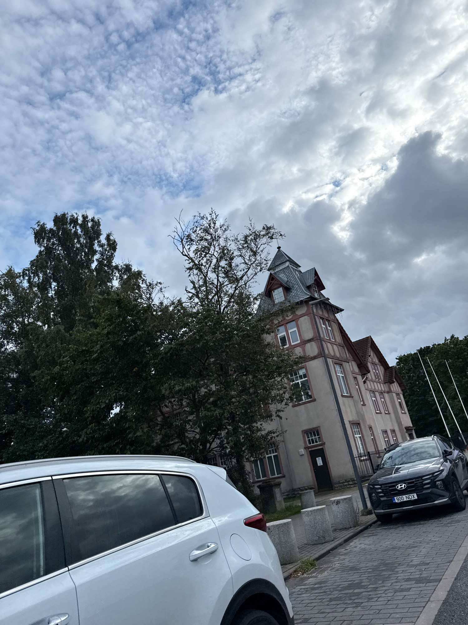 The image depicts a street scene in Tallinn, Estonia, under a cloudy sky. A light-colored car is parked in the foreground on the left, and a dark SUV is parked to its right. Behind the cars, a building with distinctive architecture, featuring a mansard roof with dormer windows and red trim, is visible. Trees with dark green foliage are situated between the cars and the building. The street is paved with cobblestones. The license plate of the dark SUV is partially visible, reading "900 NDX".