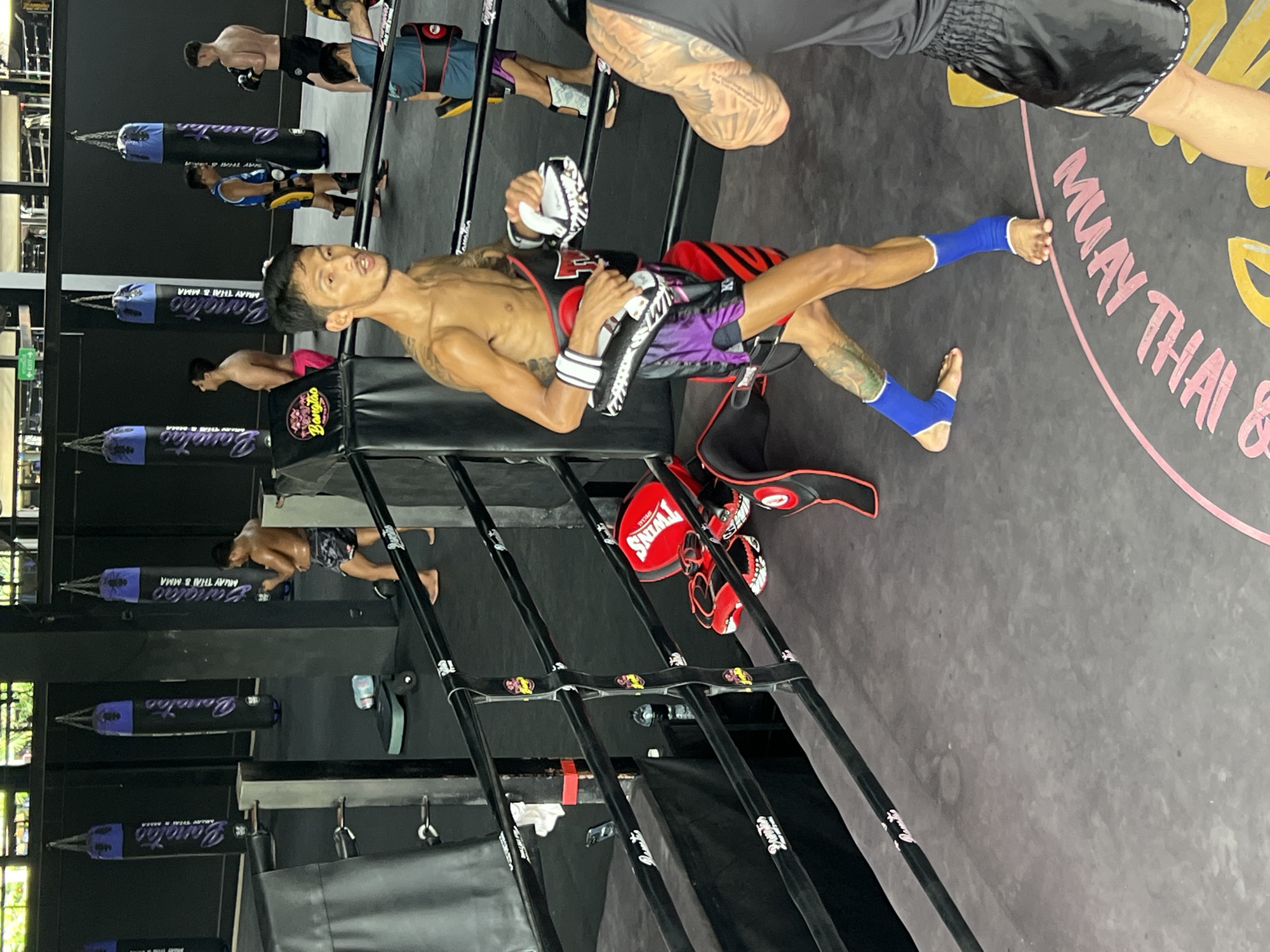 The image shows a group of people in a Muay Thai gym in Choeng Thale, Thailand. The scene is focused on two individuals in the foreground, both appearing to be male Muay Thai practitioners. One man, wearing purple and black shorts and black MMA gloves, is lying on the mat, his body positioned lengthwise. He has tattoos on his arms and legs. He is holding a pair of white and black gloves. The other man, partially visible, has blue shin guards and is also positioned on the mat.
In the background, several large, dark punching bags with purple lettering that reads "Banglao MUAY THAI & MMA" are hanging. Another person, wearing black shorts, is seen in the far background near the punching bags, appearing to be engaged in training. The gym floor is dark, with a pink circle and text that reads "MUAY THAI & MMA" visible on the mat in the lower right portion of the image. Red and black Muay Thai training equipment, including hand pads, are also visible on the mat. The overall atmosphere suggests an active training session within an indoor gym setting. The lighting appears to be artificial, typical of an indoor training facility.