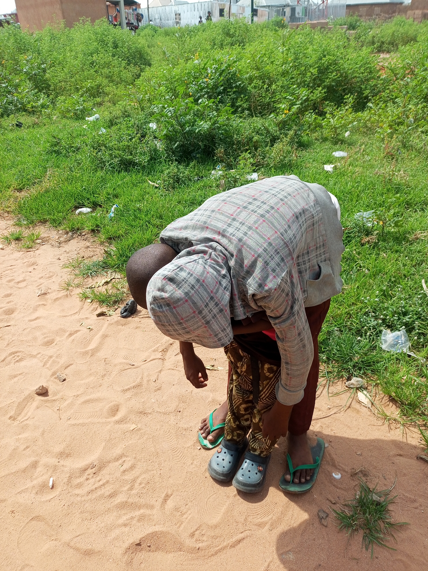 In Tempure, Nigeria, a child, viewed from behind and slightly above, is bent over with their head down, their body angled towards the ground. They are wearing a grey and red plaid long-sleeved shirt, brown pants, and a pair of dark gray clogs on their left foot and a green flip-flop on their right. The child's left hand is visible, touching their pants. They stand on a sandy surface, with a patch of green grass and sparse vegetation to their right. The background shows more overgrown greenery, with scattered white plastic debris. In the distance, a building with a brown facade is partially visible.