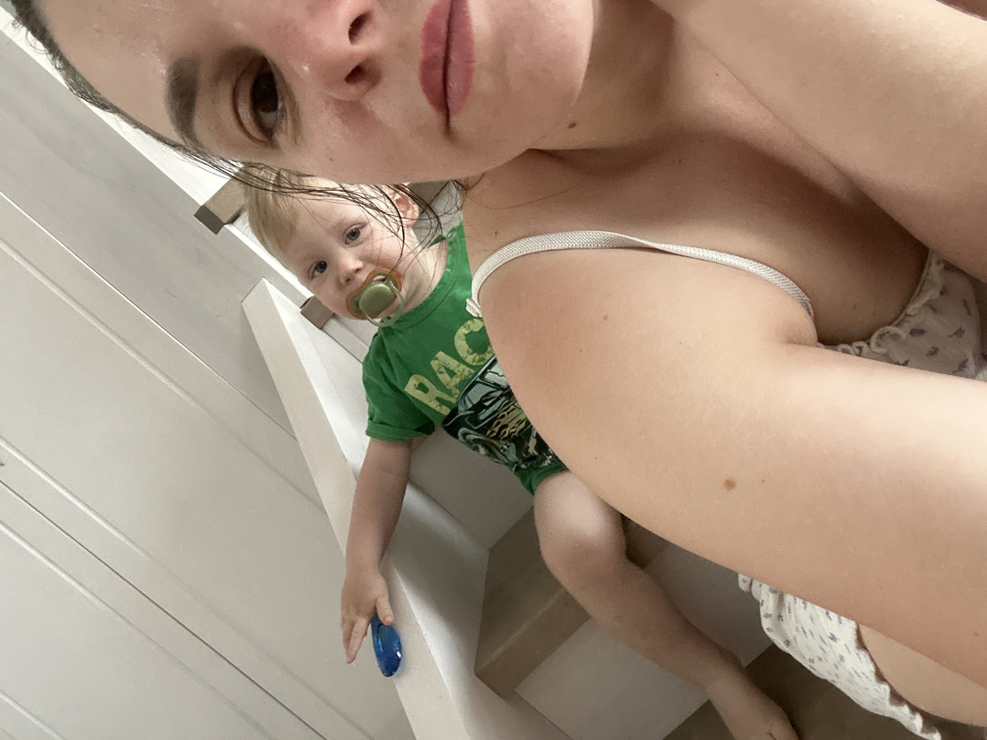 A selfie taken indoors features a woman with dark hair, light skin, and light pink lips. Her right eye is prominent in the frame. A young child with light hair is partially visible in the background, wearing a green t-shirt and holding a blue object. The child has a pacifier in their mouth. A woman’s shoulder and upper chest are also visible in the foreground, suggesting a low-angle shot. A white strap and a floral pattern on clothing are seen. Background details include a white wall with horizontal planks and a stair railing. There is no definitive information in the image to confirm the location is Chaikovsky, Russia.