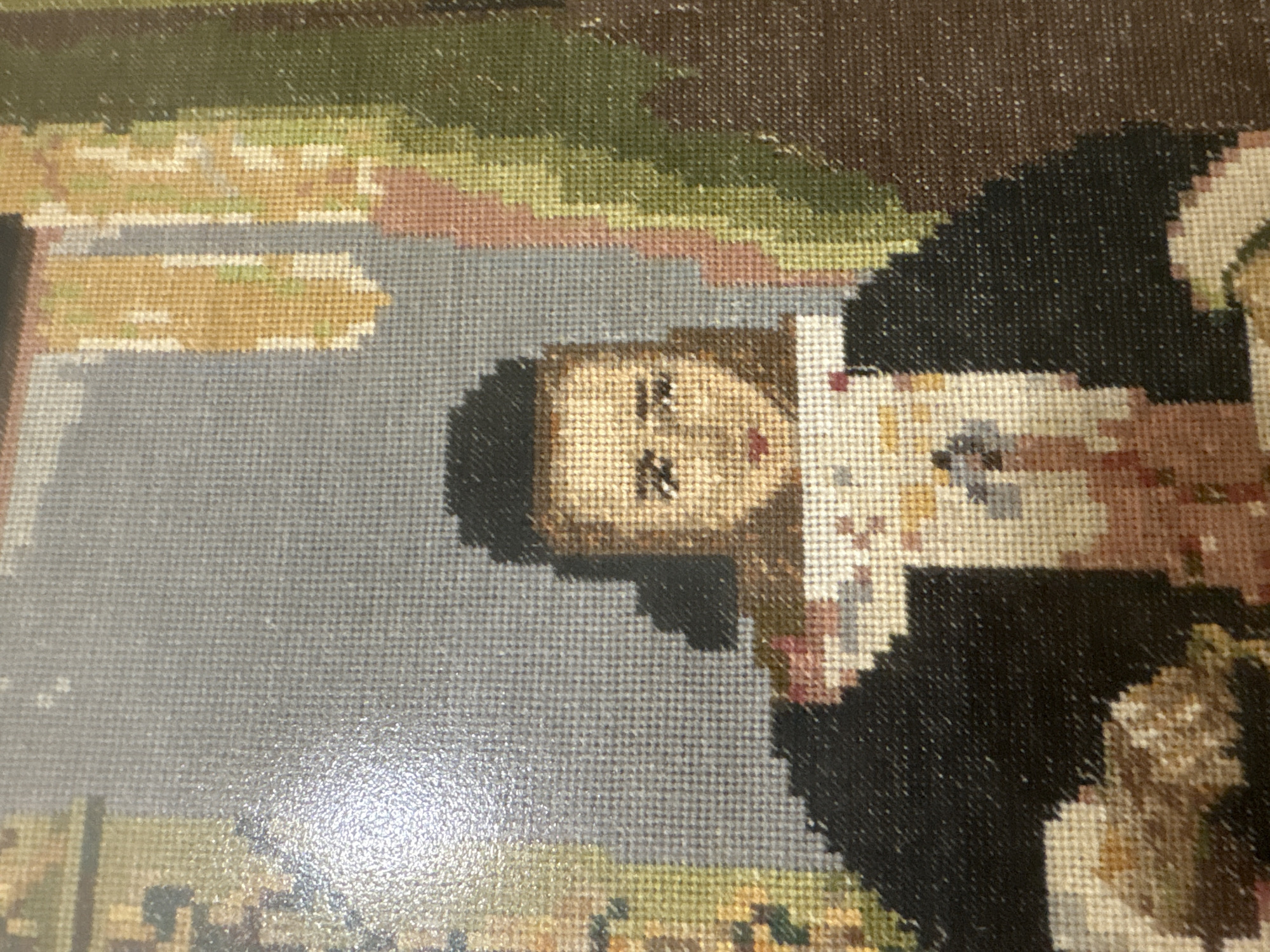 This media file is a close-up, rotated photograph of a tapestry depicting a scene with people and architectural elements. The tapestry appears to be a needlepoint or cross-stitch artwork.
The primary subject is a person's face and upper body, oriented sideways. The face has light skin tones and dark hair. Features like eyes, nose, and mouth are rendered with stitched details. The person is wearing a dark garment, possibly a dress or coat, and a white and beige patterned fabric is visible on their chest.
Behind the person, the tapestry shows a landscape with areas of green, brown, and pink, suggesting foliage or hills. There are also lighter colored areas that could represent water or paths. To the right of the person, architectural elements are depicted, including what looks like a building or structure with brown and white sections.
The image has a slightly glossy finish, which is reflecting light, particularly in the lower right portion. The texture of the tapestry, formed by individual stitches, is very apparent. The overall impression is of a traditional, possibly historical, depiction. No discernible text is visible within the tapestry. The lighting suggests it's an indoor photograph under artificial light.