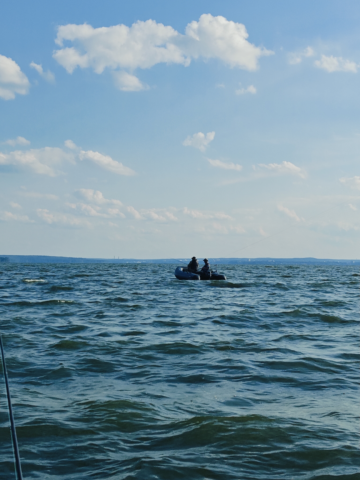 Two individuals are seated in a small, dark-colored inflatable boat on a body of water. The boat is positioned in the middle ground, with the two occupants appearing to be engaged in fishing or leisure activity. The water is choppy, with visible waves. In the background, a distant shoreline with trees and some man-made structures, possibly industrial, is visible under a bright, partly cloudy sky. The scene appears to be set on the Kama River in Chaykovsky, Russia, given the characteristic landscape and the presence of industrial elements often found in such locations. A fishing rod is visible in the foreground, extending from the lower left corner of the frame.