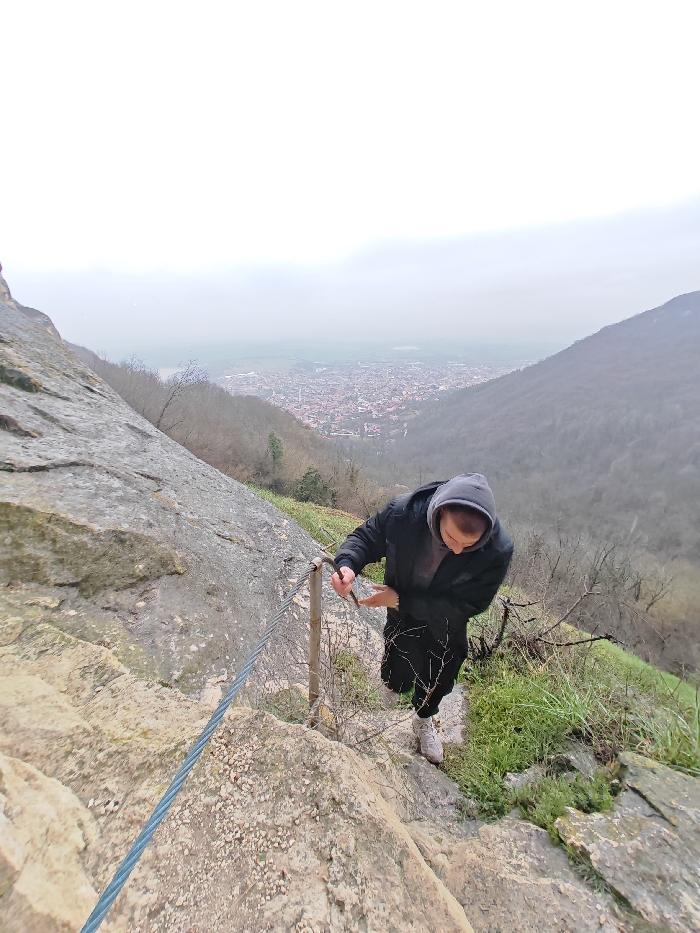 The scene depicts a steep, rocky hiking trail on a mountain overlooking the city of Shumen, Bulgaria. A single individual, appearing male, is visible in the mid-ground, ascending the path. The person is dressed in a dark grey hooded sweatshirt with the hood up, a black puffer jacket, black pants, and light-colored athletic shoes. Their right hand grasps a multi-strand metal cable, which functions as a handrail secured by metal posts embedded in the rock. The individual's head is tilted downward, focusing on the immediate path ahead. The trail consists of uneven, light-colored rock formations, interspersed with dry vegetation and patches of green grass. In the background, the urban area of Shumen is visible in a valley, characterized by numerous buildings with varying roof colors. The city is bordered by tree-covered hills, and the sky is uniformly overcast and light grey, indicating muted light conditions.
