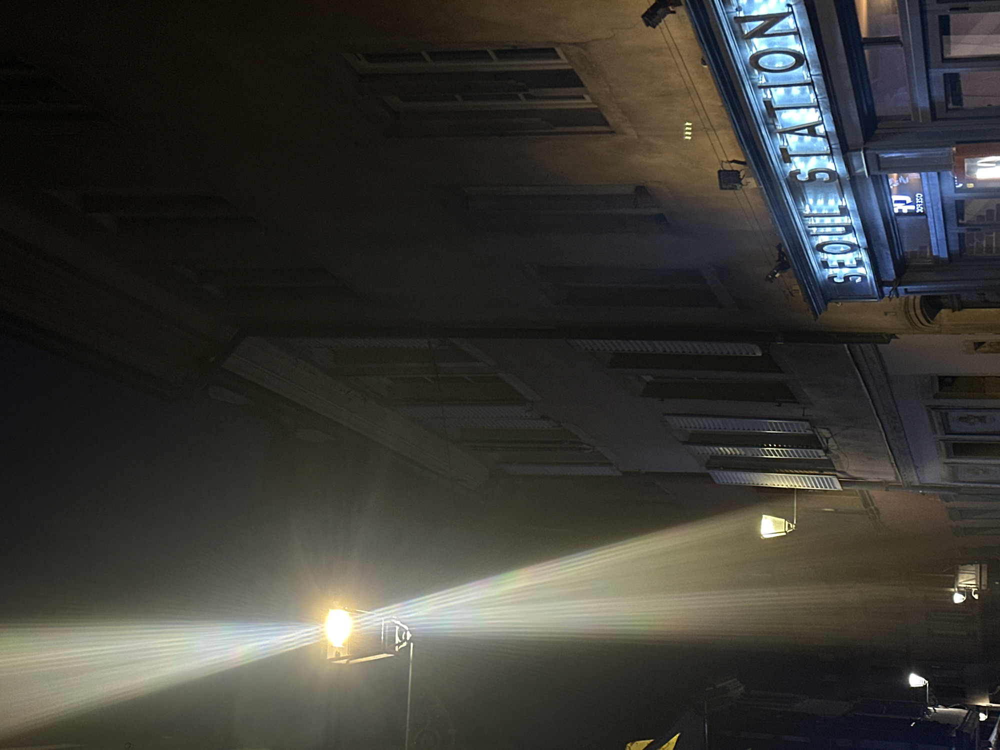The image shows the exterior of Seoul Station in Strasbourg, France at night. The building's facade is visible, with the "SEOUL STATION" sign prominently illuminated. Street lights cast bright beams of light into the scene. The architectural details of the building, including its windows and possible ventilation structures on the ceiling, are discernible. A few cars are parked on the bottom.