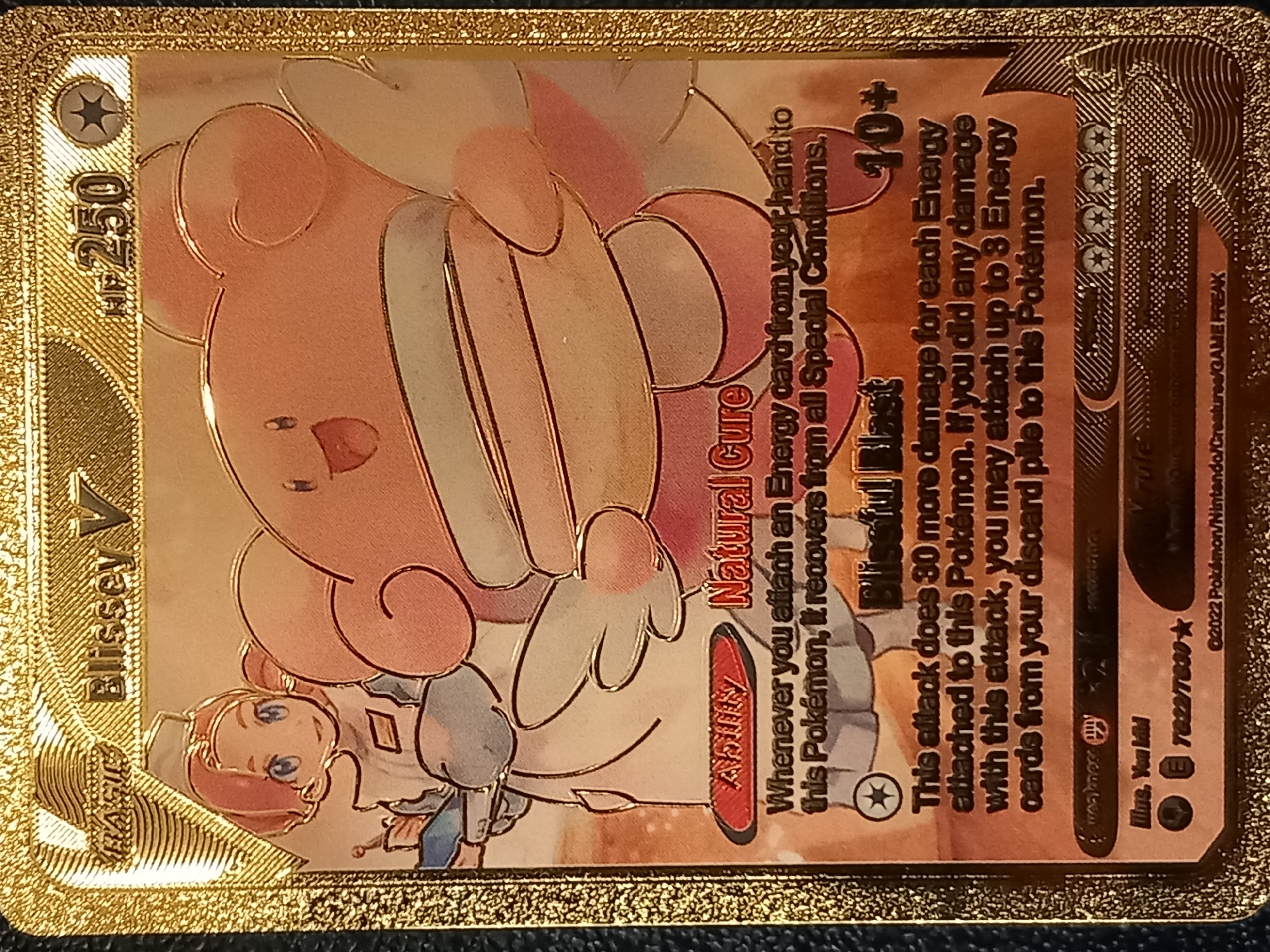 The image shows a close-up, top-down view of a gold-foil Pokemon trading card. The card features the Pokemon Blissey V, a large, round, pink creature with large, floppy ears and a contented expression. A human character, appearing to be a nurse with pink hair and blue eyes, stands to the left of Blissey. The card has text elements indicating its name, HP (250), abilities, and attack descriptions. The background of the card has a textured, patterned design in gold. The edges of the card are also gold and appear to have a raised texture. The card is placed on a dark, textured surface, possibly a table or cloth. There are no discernible environmental details related to a city.
