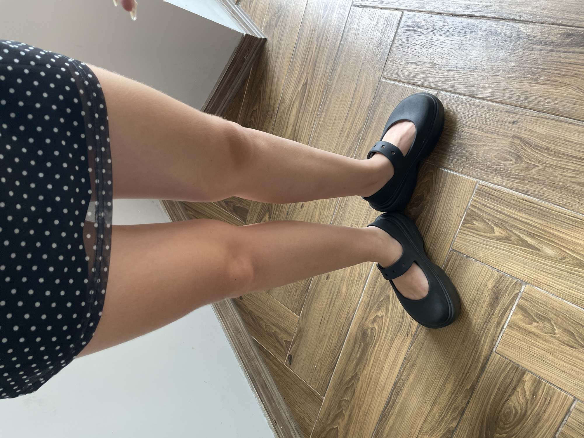 The image shows a close-up view of a person's legs and feet, standing on a wooden-tiled floor. The person is wearing a black polka-dot dress and black Mary Jane-style shoes. The background is a white wall with a wooden baseboard. There is no indication of the location being in Hanoi, Vietnam. The scene is static and focuses on the attire and footwear.