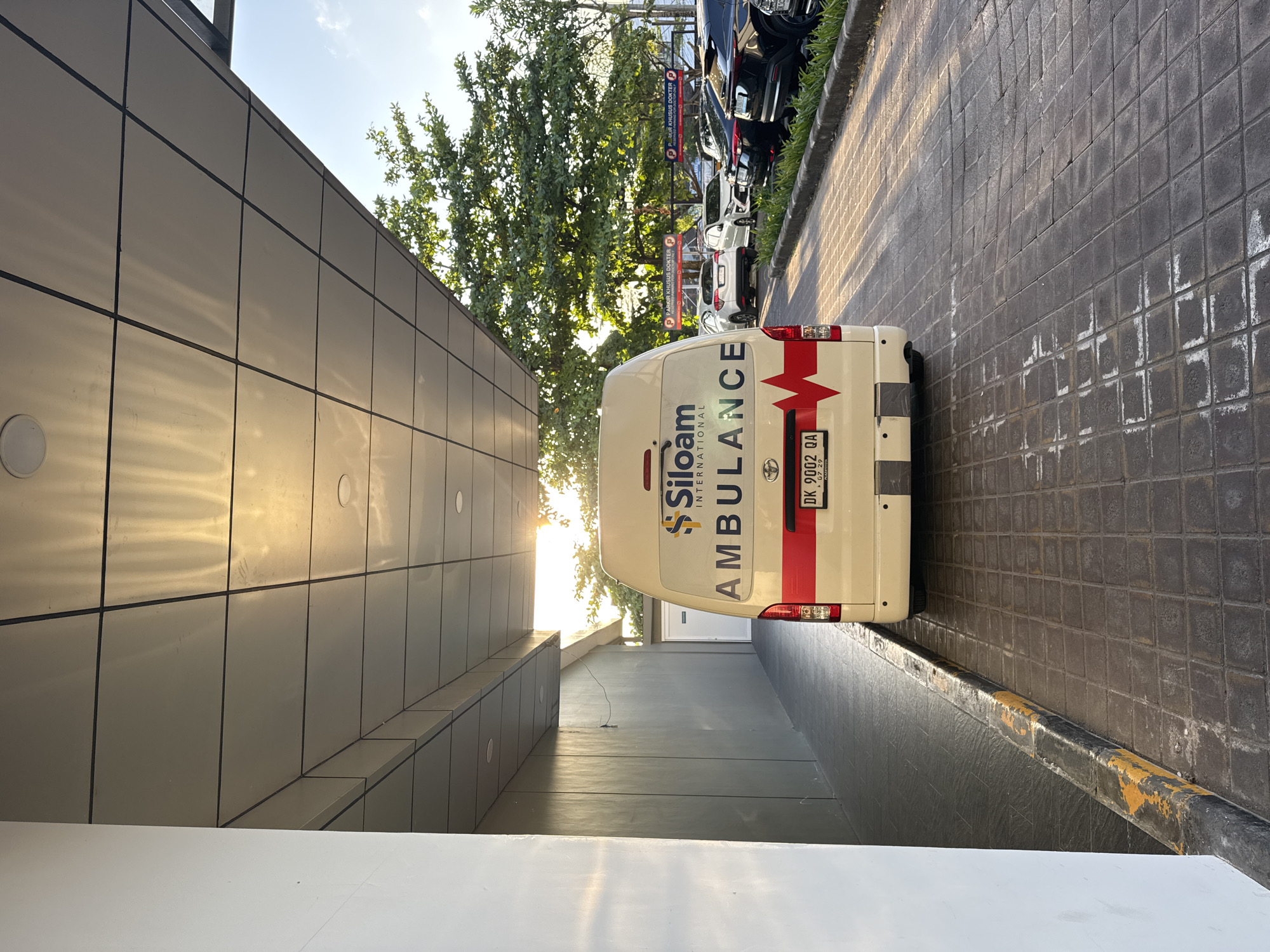 An ambulance with "Siloam International Ambulance" written on its back is parked on a paved driveway. The ambulance is white with red and white stripes on the back. A license plate with "DK 9002 QA" is visible. The ambulance is parked under a canopy with a gray tiled ceiling. To the left, a gray wall runs along the driveway. To the right, trees and parked cars are visible, with signs in Indonesian indicating "PARKIR KHUSUS DOKTER" (Special Parking for Doctors). The scene appears to be in a city, possibly Kuta, Indonesia, given the license plate format. The sun is setting, casting a warm light on the scene. There are no people visible in the image.