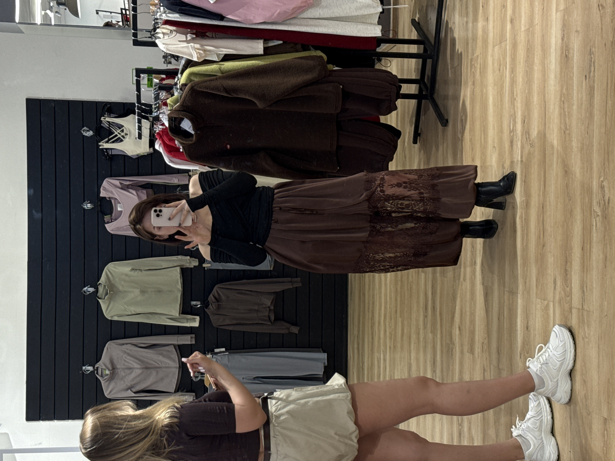 A woman takes a mirror selfie in a clothing store. She wears a black long-sleeved top and a brown, patterned maxi skirt with black boots. Another woman, wearing a brown cropped top and light shorts with white sneakers, looks at her phone near the first woman. Clothes are displayed on hangers against a dark wall and a metal rack, and the floor is wooden. There is no verifiable information to confirm that this image was taken in Perm, Russia.