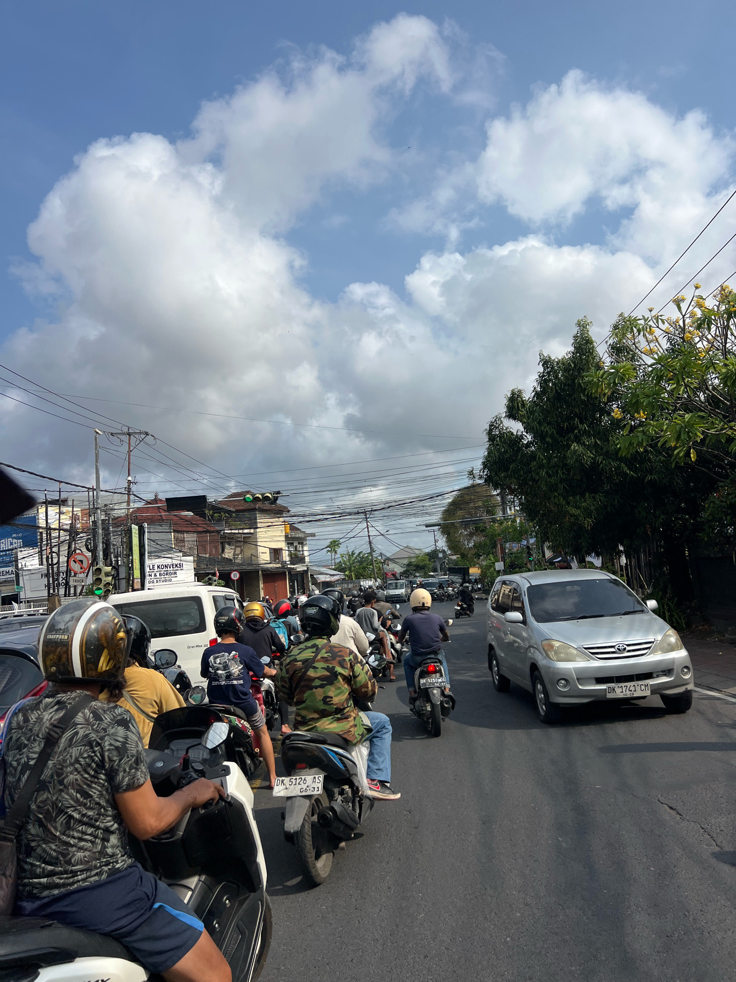 This is a street-level, medium shot captured on a sunny day in Denpasar, Indonesia, depicting moderate traffic congestion. The foreground features a person riding a white scooter, wearing a black and white patterned shirt and dark blue shorts, and a helmet with gold and white accents. Behind them, other motorcyclists are visible, many wearing helmets and casual clothing, including a person in a camouflage-patterned shirt. A silver minivan is prominently positioned in the middle of the road. The background reveals a street lined with buildings, utility poles with numerous wires, and trees with green foliage and yellow flowers. Traffic signals are visible, one displaying a green light. The sky is blue with scattered white clouds. The road surface is asphalt.
