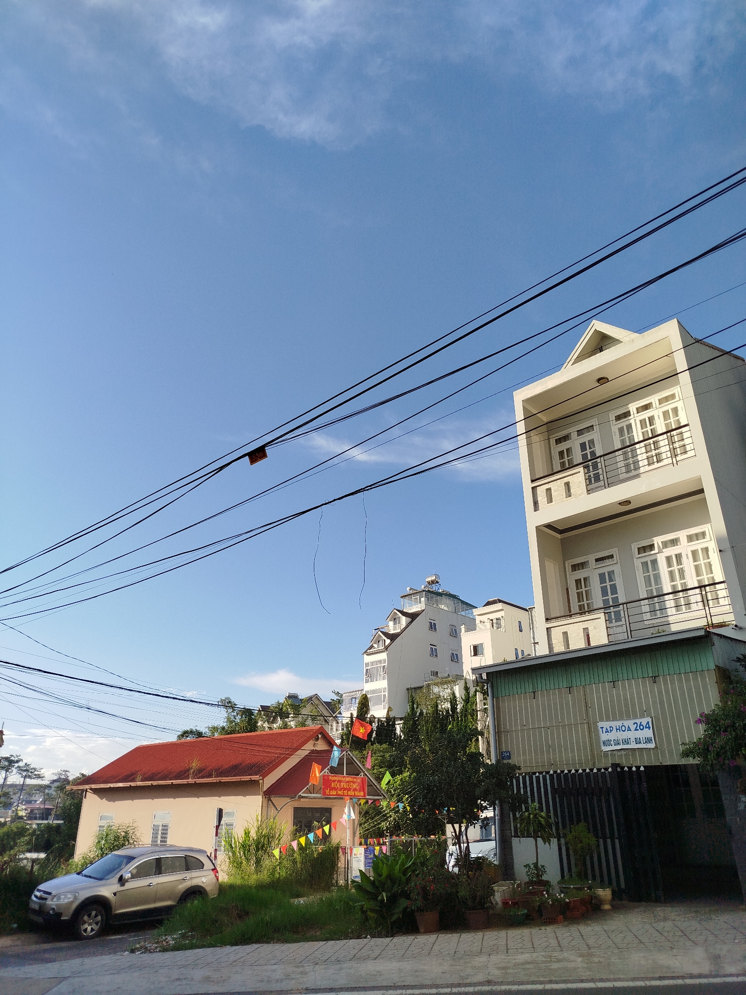 This is a daylight, eye-level, outdoor, medium shot of a street in Da Lat, Vietnam, with a focus on residential buildings and overhead electrical wires. The sky is a clear, bright blue with some wispy clouds. In the foreground, a grey SUV is parked on the left side of the frame. Next to it is a building with a reddish-orange, gabled roof. This building has several windows and is decorated with colorful pennant banners and Vietnamese flags. A sign on this building reads "HOI TRUONG CO DAN PHO HIEN THANH".
To the right of this building is a taller, modern, multi-story white and grey residential building with balconies and multiple white-framed windows. Further back and to the left, other white multi-story buildings are visible, some with mansard roofs and balconies. One of these buildings has a solar water heater on its rooftop. In the mid-ground, a long, narrow building with a corrugated metal facade and a green awning is situated. A sign on this building displays "TAP HOA 264 NGUOC GIAI KHAT - BIA LANH".
Numerous black electrical wires crisscross the sky, obscuring parts of the buildings and sky. The ground in front of the buildings is paved with grey interlocking stones. Lush green vegetation, including trees and potted plants, is visible around the buildings. The overall impression is of a residential street in a Vietnamese city, likely on a sunny day. The presence of multiple flags suggests a local gathering or event.