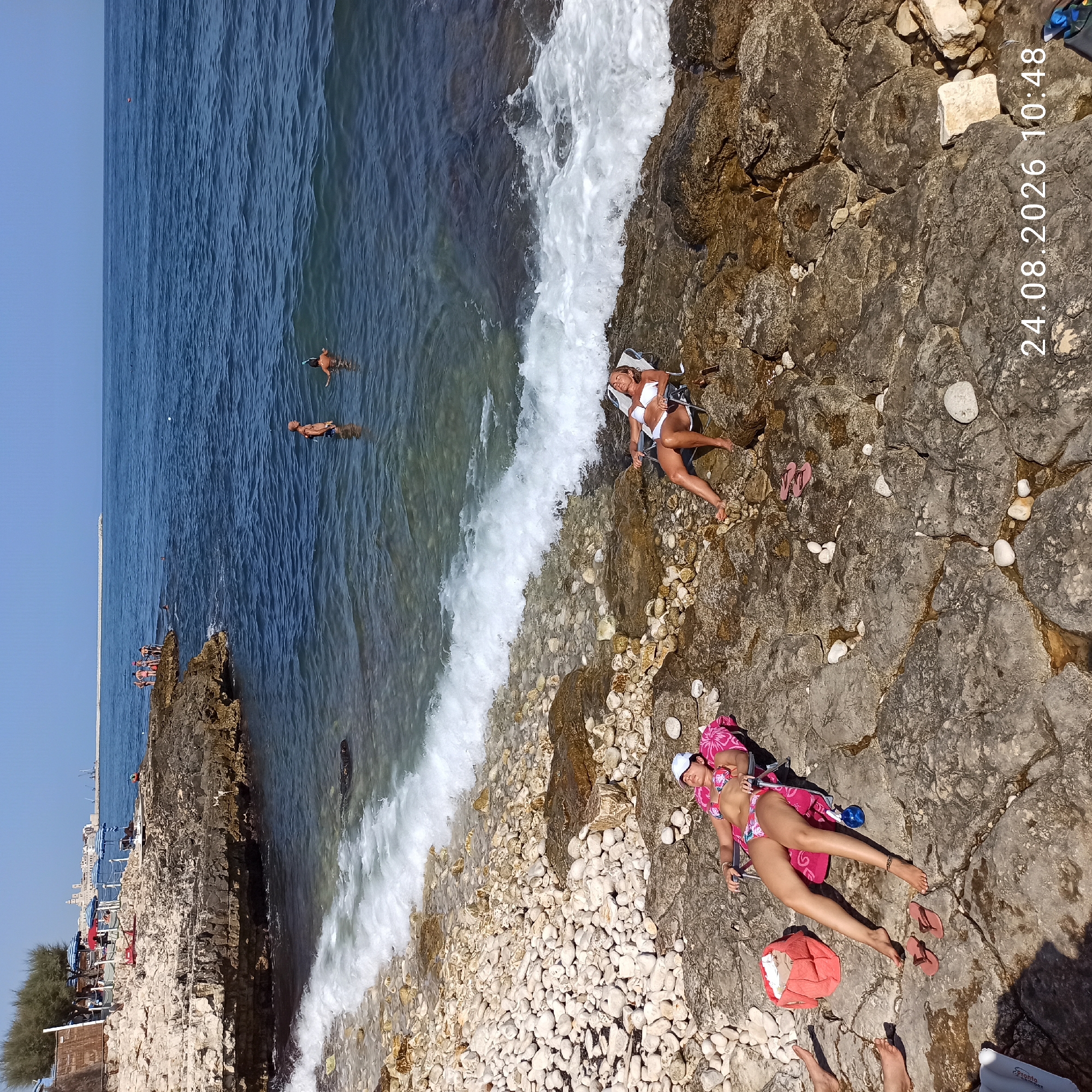 The image depicts a rocky coastline near Molfetta, Italy, on August 24, 2026, at 10:48 AM. The sea is a deep blue, with waves breaking on the shore. Two women are sunbathing on chairs on the rocky beach. One woman, wearing a floral bikini and a white cap, is lying on a pink towel. The other woman, in a white bikini, is also reclining on a chair. Several people are visible in the water in the distance, some swimming and one snorkeling. A small village with white buildings and a church steeple can be seen on the distant coast, suggesting a coastal urban setting. The beach is made up of smooth white pebbles and larger, rougher rocks. Personal items such as a coral-colored bag and flip-flops are scattered on the rocks.
