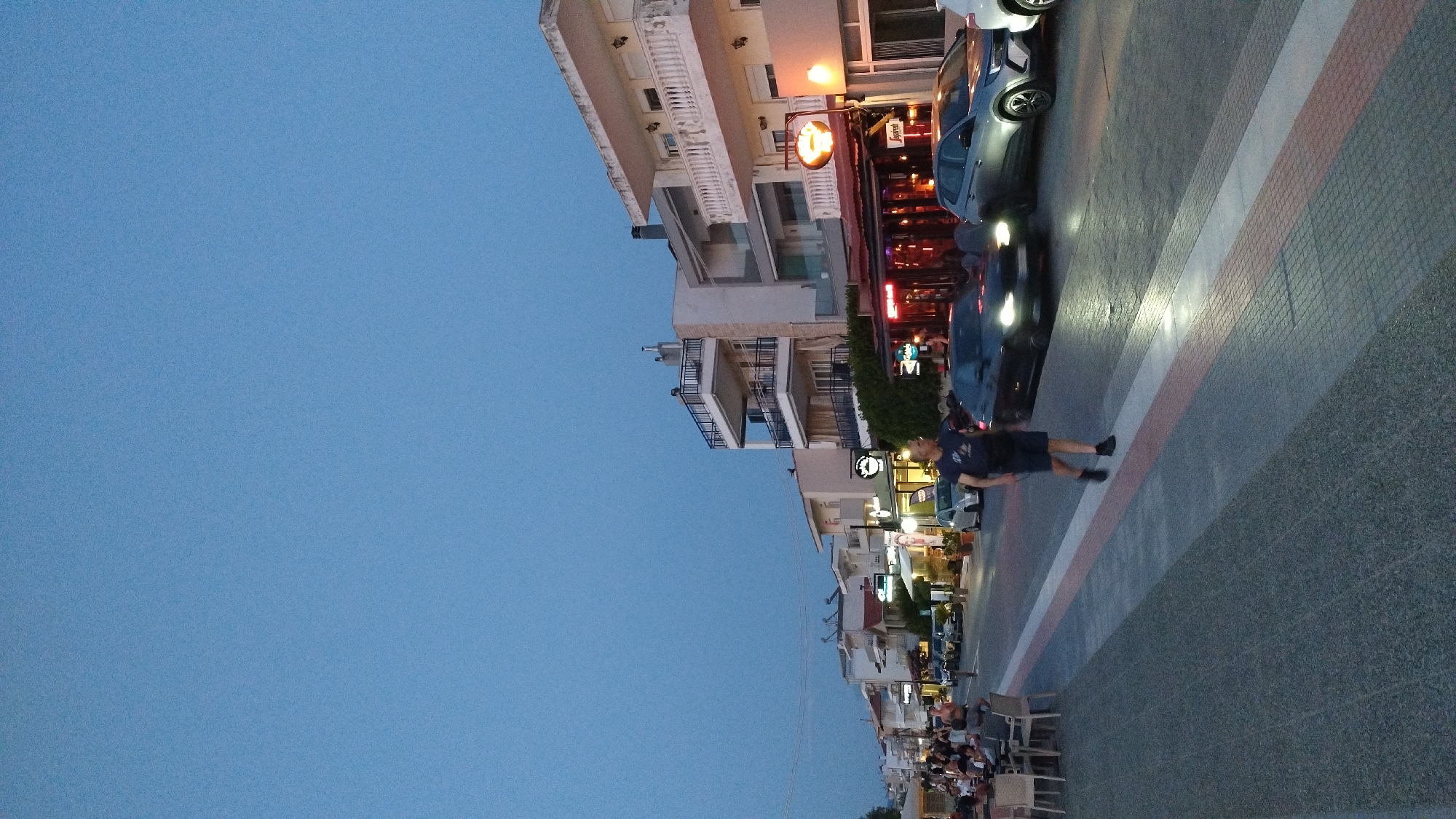 The image captures a street scene at dusk in Neoi Epivates, Greece. A man in a dark t-shirt and shorts is walking across a crosswalk on the right side of the image. Cars are driving on the street, with headlights illuminated. On the left side of the street, there are tables and chairs set up outside a restaurant or cafe, with people seated. Buildings with multiple balconies line the street. The sky is a clear, dusky blue. The overall atmosphere appears to be relaxed, with the beginning of evening activity on the street.