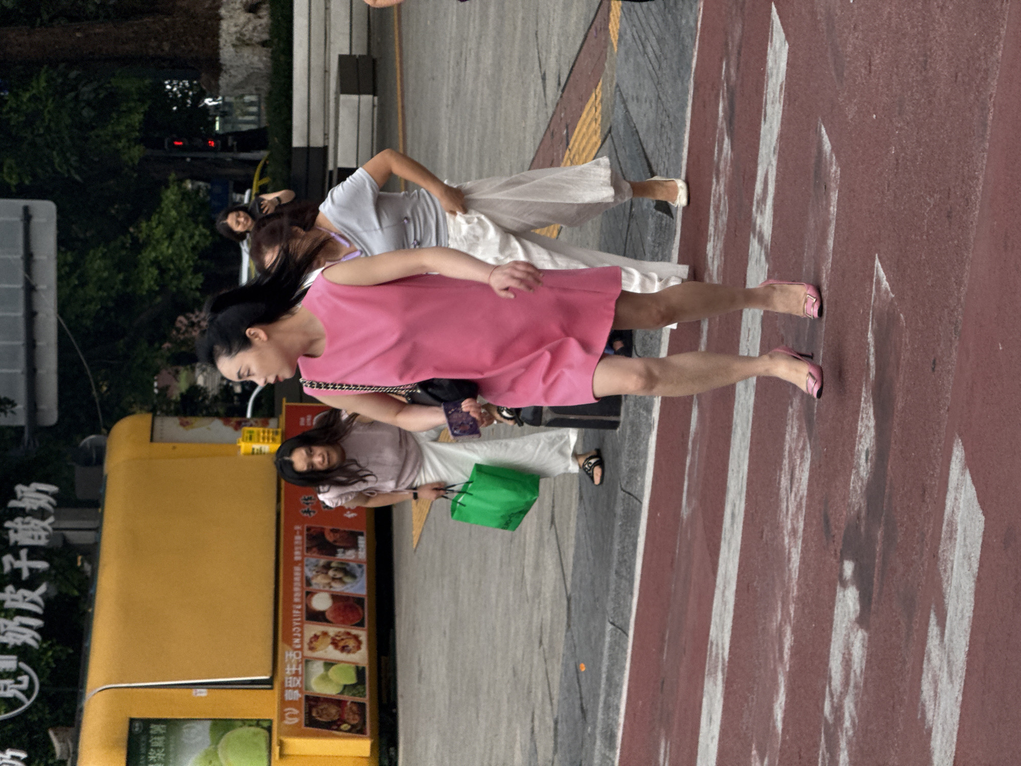 In Yuzhong District, China, a group of individuals are crossing a red and white striped crosswalk. A woman in a pink sleeveless dress and pink high heels is in the foreground, looking to her left. Behind her, another woman in a light gray t-shirt and loose white pants, wearing black sandals, is carrying a green shopping bag in her right hand and looking down. Further behind, a third person, partially visible, wears light gray pants and white shoes. The background includes a yellow food cart with various food images displayed on its side, greenery, and trees. The ground surface is wet, suggesting recent rain.