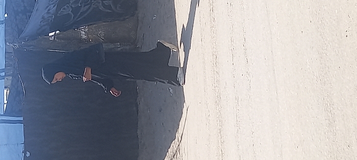 The image depicts a single individual walking on a light-colored, unpaved surface in Khan Yunis, Palestinian Territory. The person is dressed in a black full-length garment, consistent with an abaya, and a black head covering, with white athletic shoes visible on their feet. The individual is captured mid-stride, moving from the right side of the frame towards the left. Strong shadows are cast on the ground, indicating direct sunlight. In the background, behind the person, makeshift structures constructed from dark tarpaulins and fabric are visible, suggesting a camp or informal settlement environment. A segment of a blue tarp is visible at the top left of the background structures.