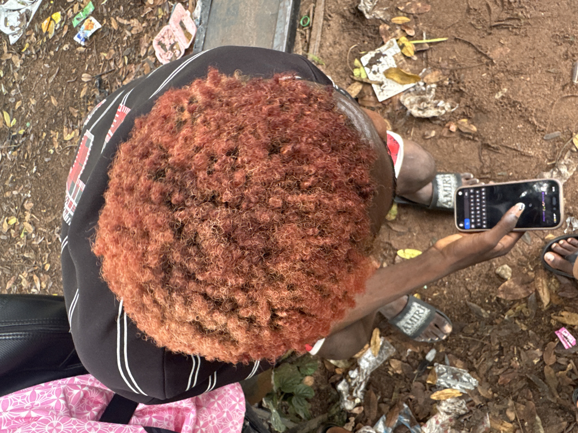 The image shows a person from a top-down perspective, focusing on their head and upper body. The person has bright orange, curly hair and is wearing a black shirt with white stripes and red accents. They are holding a smartphone in their right hand, and their left hand is partially visible. The person is wearing sandals. The background consists of dirt, fallen leaves, and scattered trash, suggesting an outdoor environment. There is also a pink and white patterned fabric visible on the left side of the image, and part of a black bag or backpack. The ground appears to be a mix of soil and debris. The scene is located in Ihiagwa, Nigeria.