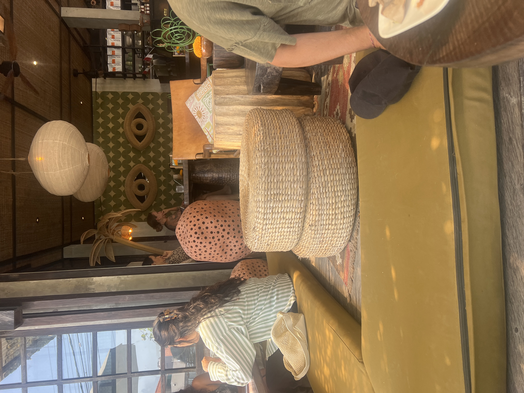 In a sun-drenched interior, possibly a cafe or restaurant in Mengwi, Indonesia, a woman with long, dark hair tied back with a claw clip is seated at a wooden table, wearing a green and white striped long-sleeved shirt. To her left, a straw bag rests on a mustard-yellow cushioned bench. In the background, a large, salmon-colored, perforated decorative object is visible, along with a woven straw palm tree accent lamp and a wall adorned with a green, diamond-patterned wallpaper featuring two large, woven eye motifs. Two stacked, round, woven straw ottomans are placed on the floor. To the right, a person wearing a light green t-shirt is partially visible, seated at a dark wooden table with a white plate holding food. The environment suggests a bohemian or eclectic design aesthetic, with natural materials and warm lighting.