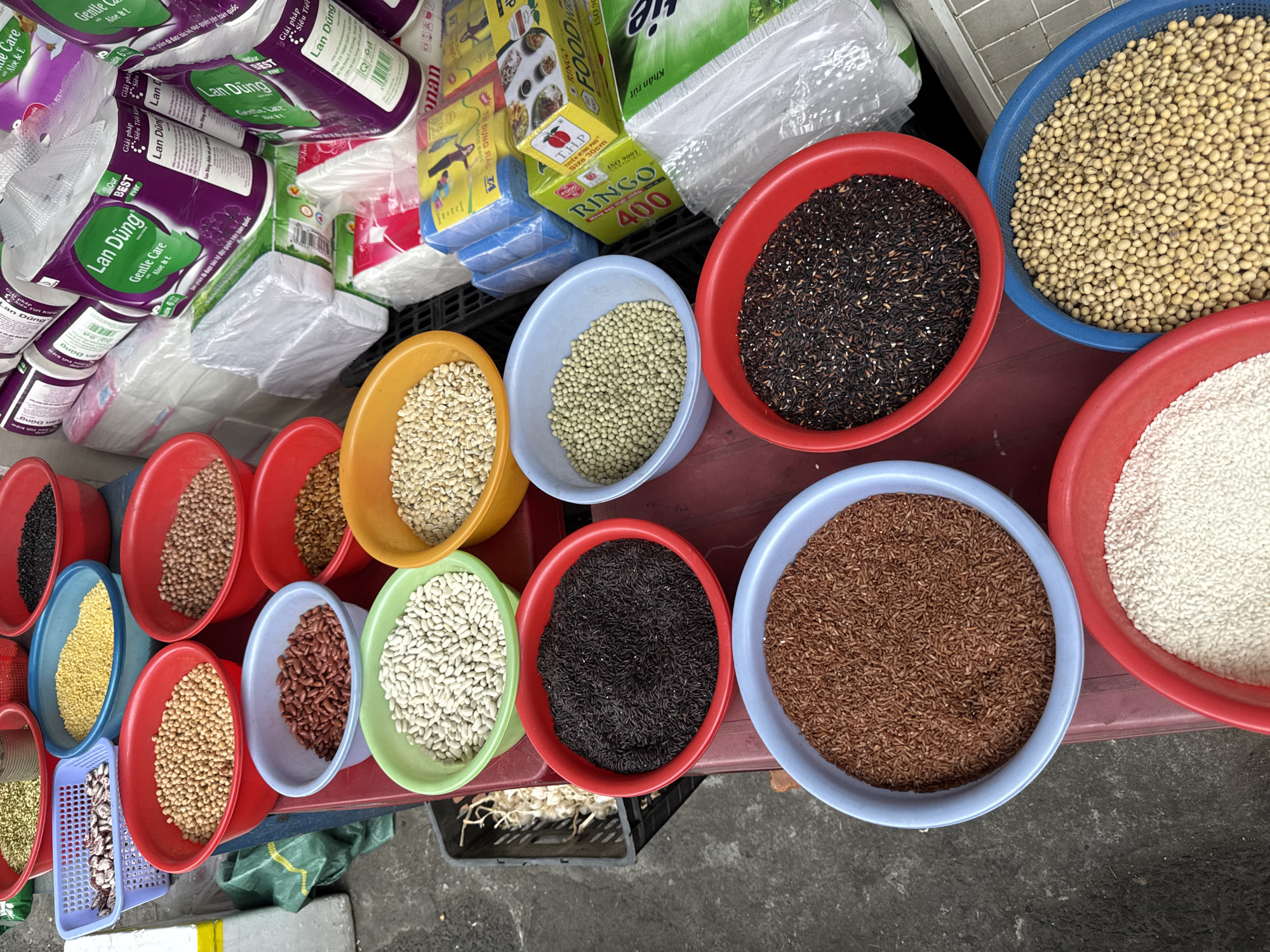This is a street market scene in Đà Nẵng, Vietnam, showing a vendor's table laden with various grains and beans. Several colorful plastic bowls and baskets are arranged on a red tabletop. The bowls contain different types of grains and legumes, including soybeans, lentils, rice, and beans of various colors (white, brown, red, and black). The table also features stacks of branded products, including Lan Dũng Gentle Care toilet paper, Ringo food wrap, and T.H.P. Túi Đựng Rác plastic bags. A black plastic crate is visible beneath the table. The table appears to be set up on a sidewalk or street corner.