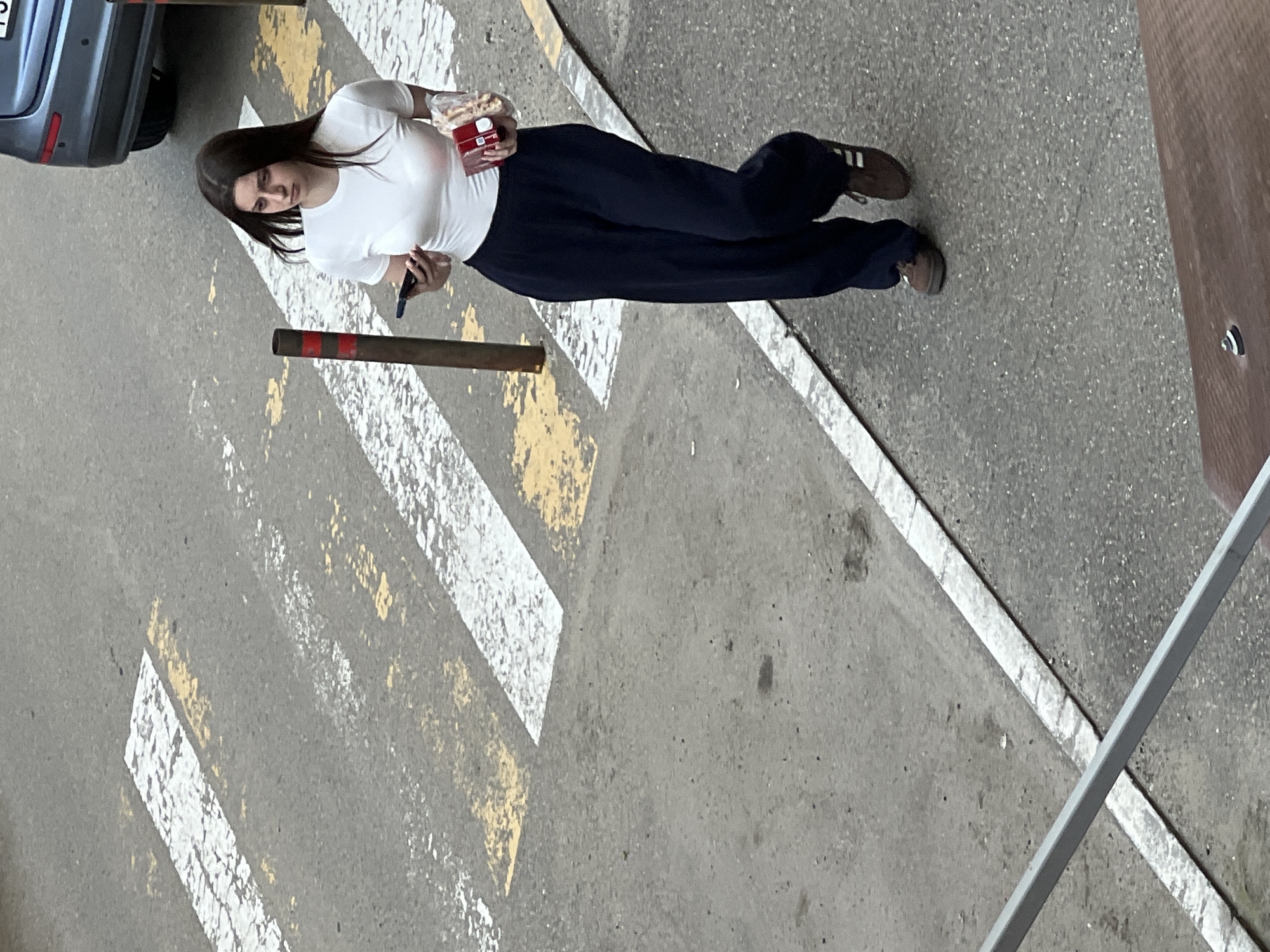 An elevated shot captures a young woman standing on an asphalt surface in an urban environment in Krasnodar, Russia. She has long, dark hair and is wearing a white short-sleeved t-shirt, dark blue wide-legged trousers, and brown sneakers with white stripes.
In her right hand, she holds a clear plastic bag filled with light-colored snacks, possibly breadsticks, along with a small red and white box that resembles a cigarette pack. Her left hand holds a dark smartphone. Her gaze is directed slightly to her left, with a neutral and observant expression.
The ground is marked with worn white and yellow painted lines, characteristic of a pedestrian crossing or parking zone. A grey car is partially visible, parked on the left side of the frame behind her. Two rusty metal bollards with faded red and white bands are positioned in the foreground. On the far right, a brown, textured surface, possibly a building or fence, is partially visible. The lighting suggests daytime, with diffused light indicating an overcast sky. No discernible text is visible on any items or surfaces.