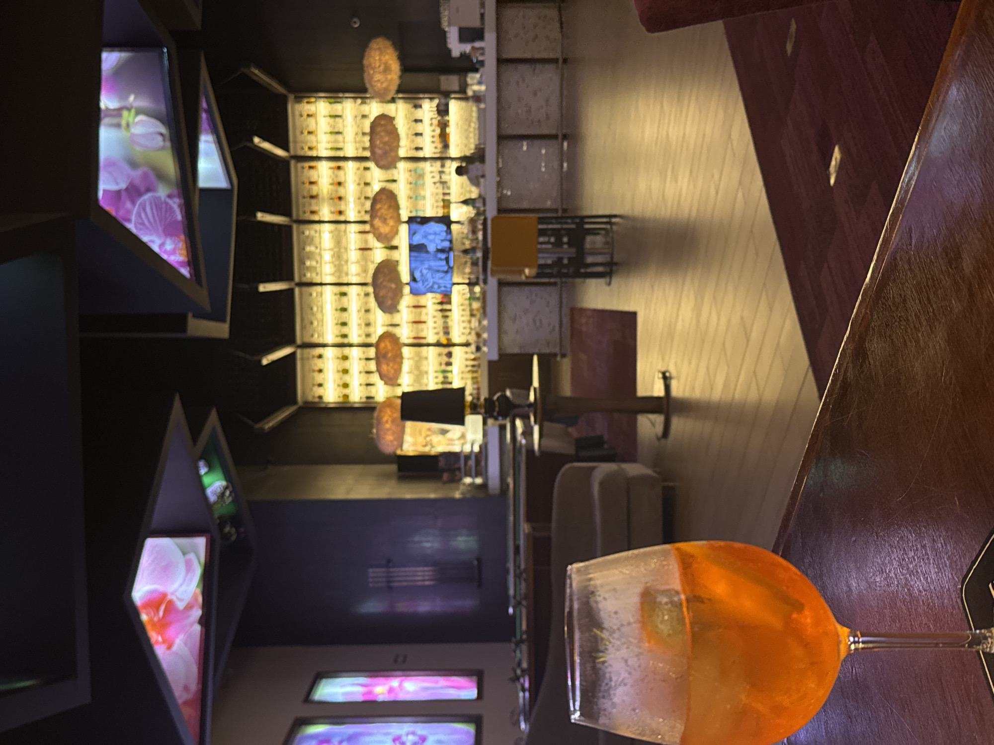 The photograph depicts an interior setting, likely a bar or upscale restaurant in Playa del Carmen, Mexico, as indicated by the type of establishment and possible geographical location inferred from the scene. The foreground features a partly consumed, orange-colored cocktail in a stemmed glass, resting on a dark wooden table. The middle ground reveals a seating area with upholstered chairs and tables, transitioning into a bar area. The bar is well-stocked with various bottles, illuminated by decorative pendant lights resembling spheres. In the background, a wall displays what appears to be digital art installations or screens with colorful floral imagery. The environment suggests a modern, stylish atmosphere with a focus on visual aesthetics. The lighting is subdued, creating a warm ambiance within the space.