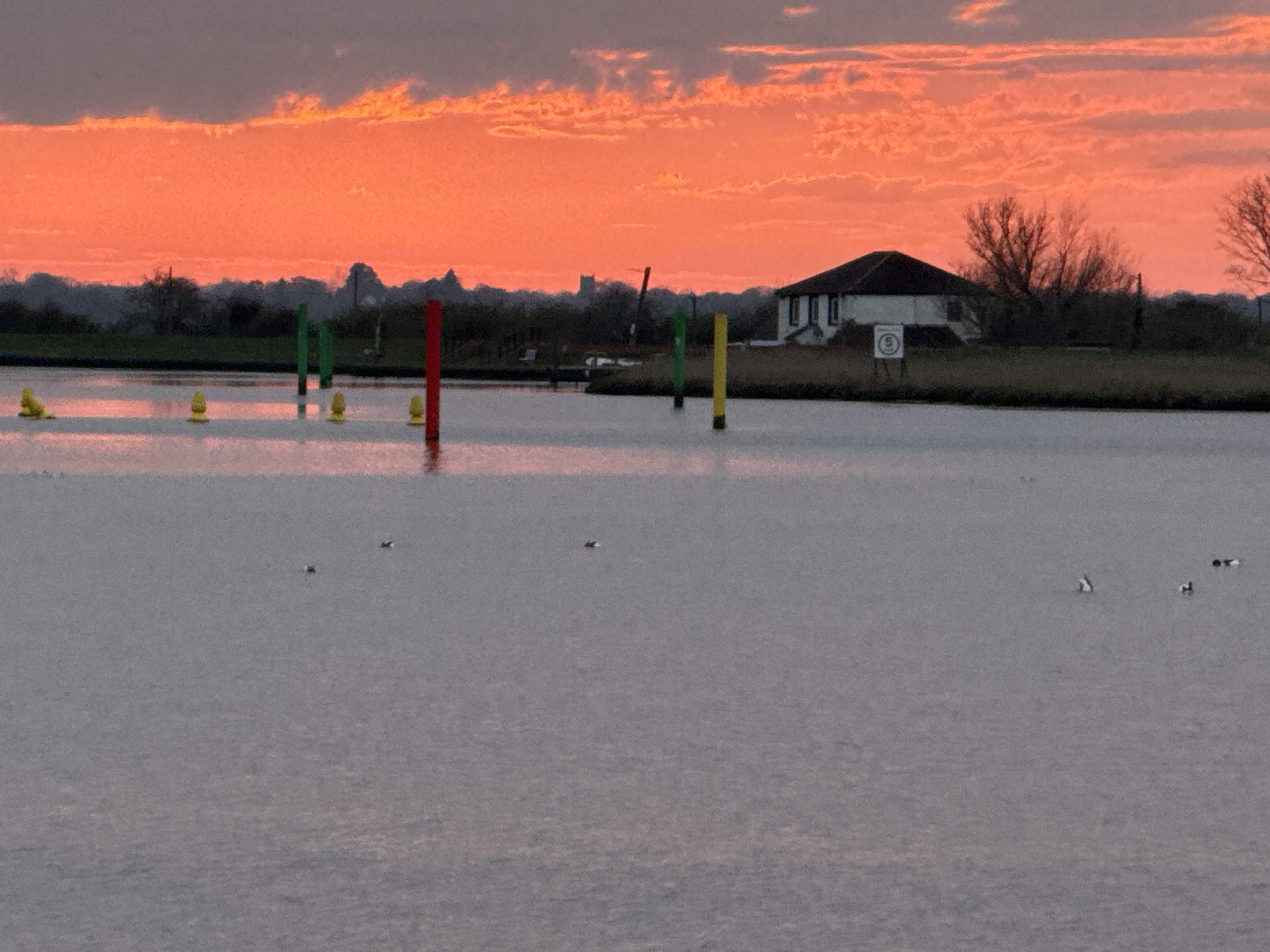 The image depicts a tranquil scene at sunset, likely in Great Yarmouth, United Kingdom. A body of water, possibly the River Bure, stretches across the foreground. Several colorful buoys and posts are positioned in the water, indicating channel markers. On the distant shore, a white house with a dark roof is visible. A speed limit sign is also present near the house. Beyond the shoreline are trees and buildings silhouetted against the fiery orange sky, suggesting either sunrise or sunset. Several birds can be seen on the water.