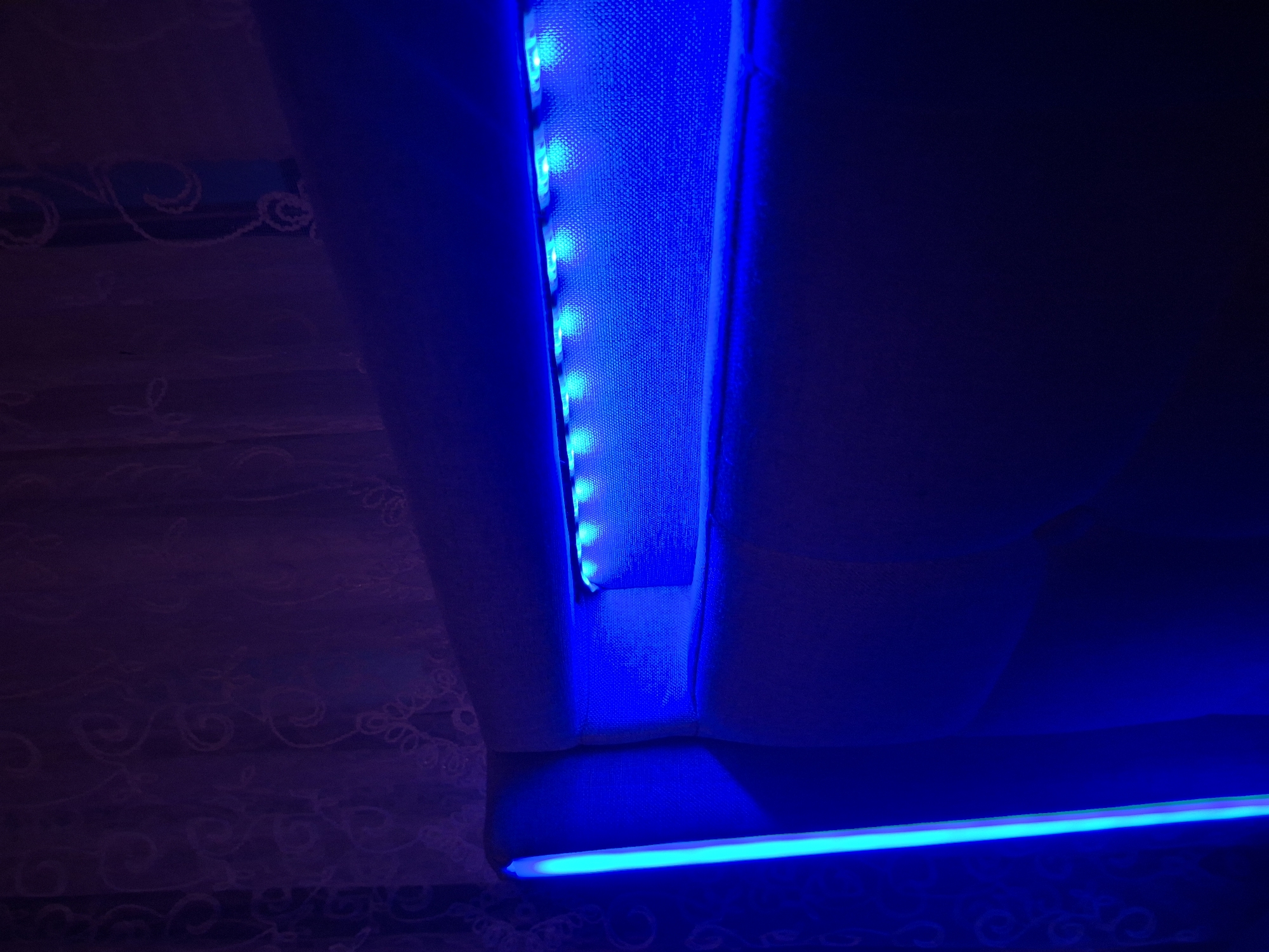 The image shows a close-up view of a headboard with integrated blue LED lighting. The headboard appears to be upholstered in a light-colored fabric. A vertical strip of bright blue light is visible on the left side of the headboard, and a horizontal strip of blue LEDs illuminates a recessed area within the headboard. The background consists of dark, possibly purple-tinted curtains with a subtle patterned texture. The lighting creates a moody and atmospheric effect. The image is taken indoors, likely in a bedroom. There is no indication of a specific location within the city of Evere, Belgium.