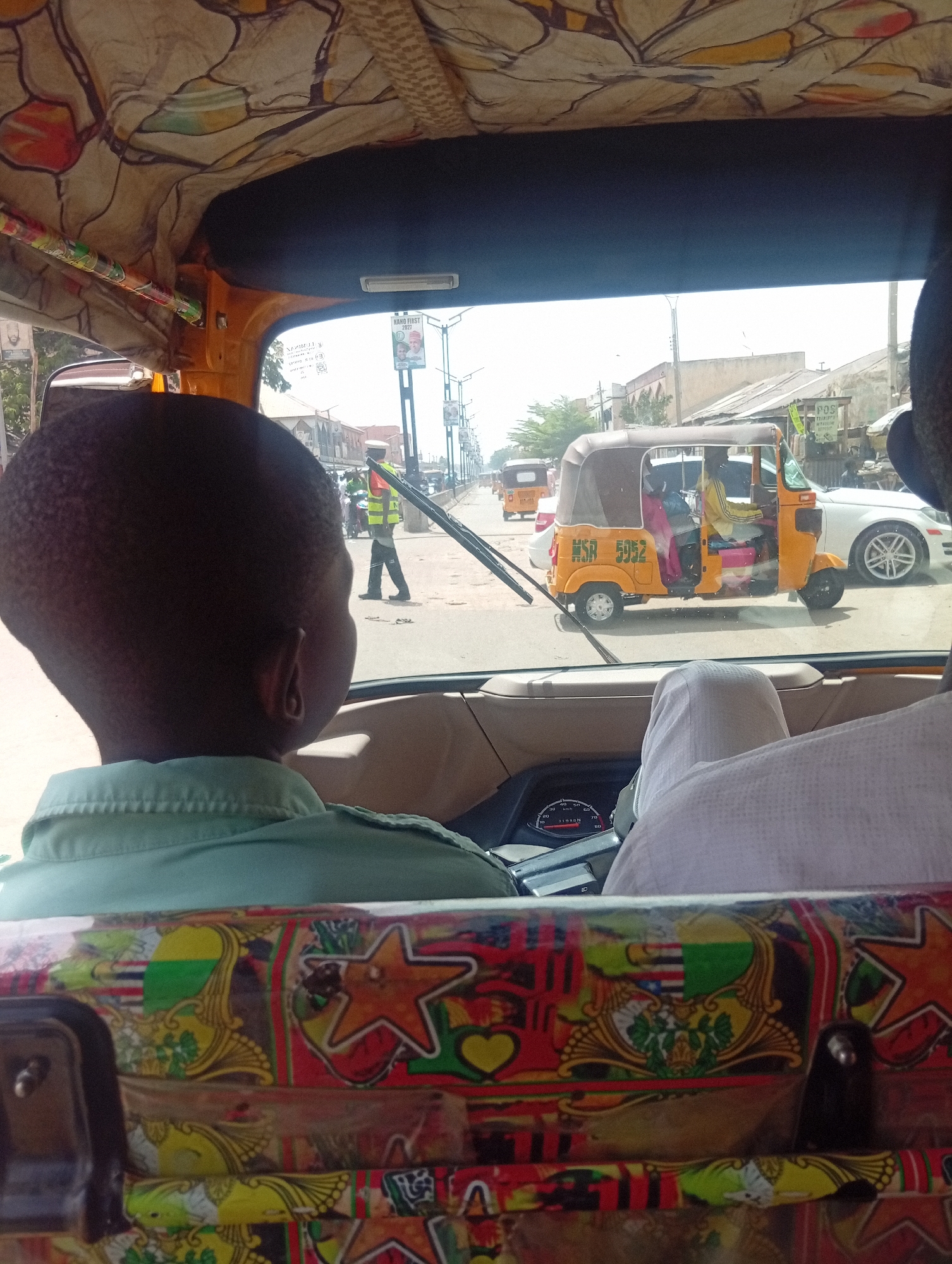 From within a vehicle, likely a tuk-tuk, a daytime street scene unfolds in Gama, Nigeria. Outside, a traffic warden wearing a reflective vest stands near another tuk-tuk bearing the license plate "MSA 5952". A white car is parked nearby. Inside the tuk-tuk from which the photo is taken, two individuals are visible; a person in a green shirt in the foreground and another person in a white shirt to the right. The interior of the vehicle is decorated with patterned fabric. A speedometer is visible on the dashboard.