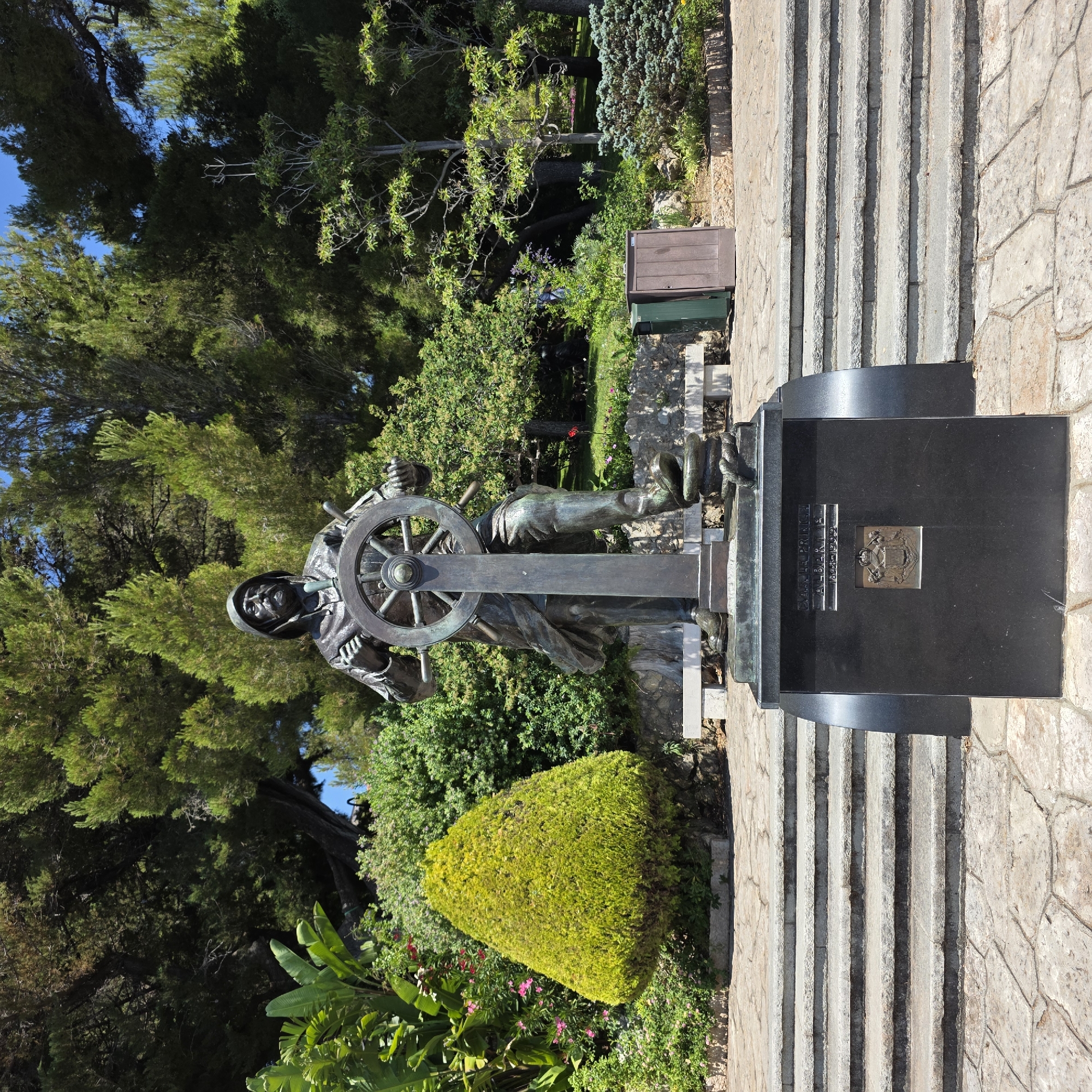 This media file captures a statue of a sailor at a ship's helm, set against a backdrop of lush greenery and a bright blue sky. The statue, made of weathered bronze, depicts a man wearing a hat and coat, his hands firmly on the wheel. Behind him, a dark granite monument stands tall, bearing an inscription and a coat of arms. The monument has a plaque with an emblem and text that reads "SON ALTESSE SÉRÉNISSIME LE PRINCE ALBERT Ier 1848-1922".
The setting appears to be a memorial or park, with stone steps leading up to the monument and statue. The surrounding vegetation is vibrant, with a mix of trees, shrubs, and flowering plants. The sunlight suggests it is daytime, and the clear sky indicates fair weather. The overall mood is one of remembrance and respect, honoring Prince Albert I. The location is identified as Monaco.