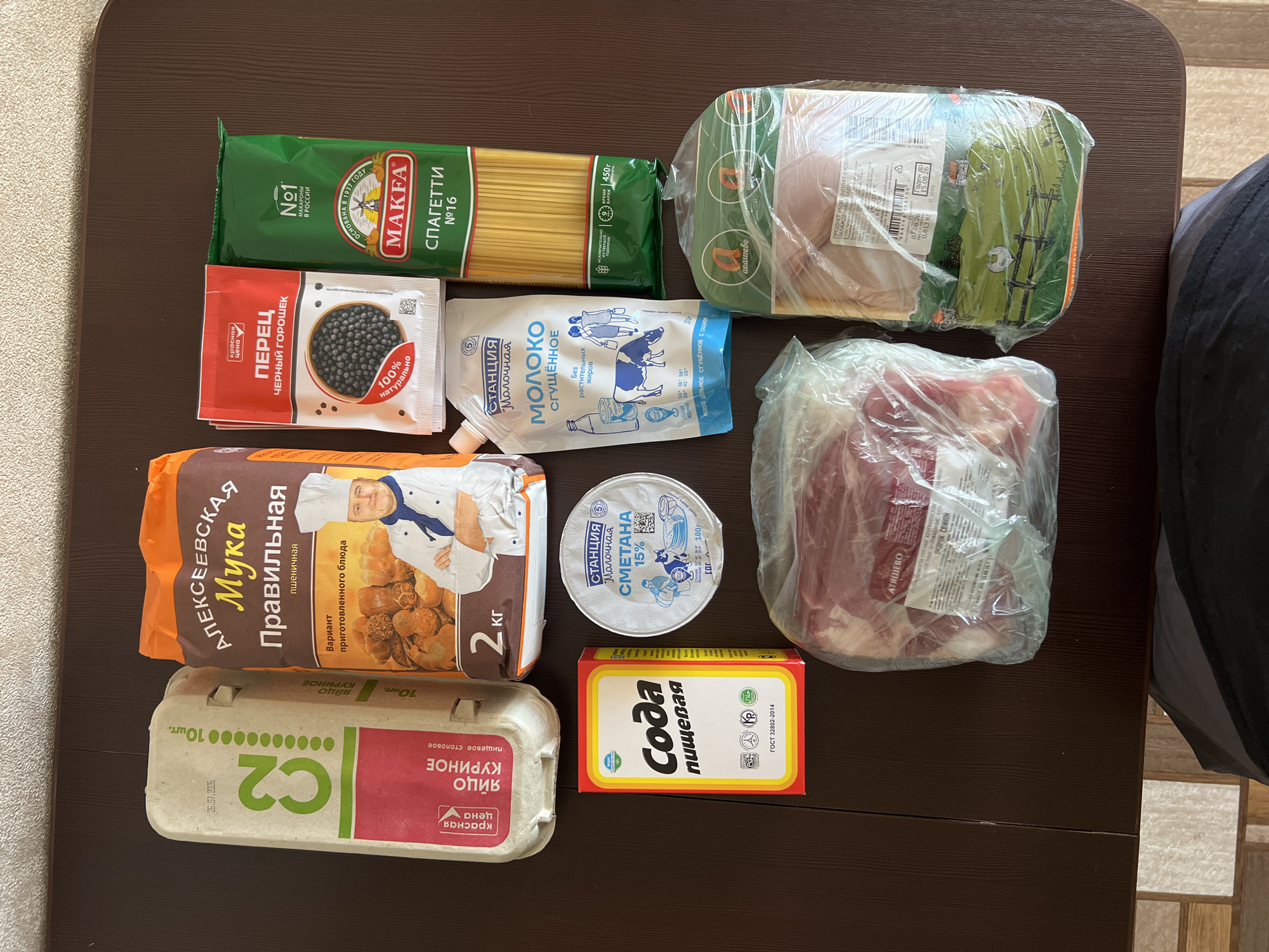 This overhead shot displays a collection of groceries laid out on a brown, wood-grain textured surface, possibly a table. The products include a carton of eggs labeled "C2" and "ЯЙЦО КУРИНОЕ" (chicken egg), a 2kg bag of flour named "АЛЕКСЕЕВСКАЯ Мука Правильная" (Alexeevskaya Flour Proper), a small red and yellow box of baking soda "Сода пищевая" (Baking Soda), a white pouch of condensed milk "МОЛОКО СГУЩЕННОЕ" (Condensed Milk), a green package of "MAKFA" spaghetti, a red sachet of black peppercorns labeled "ПЕРЕЦ черный горошек" (Black Peppercorns), a tub of sour cream labeled "СМЕТАНА 15%" (Sour Cream 15%), a package of raw pork, and a package of raw chicken. The background appears to be a neutral-colored wall with a subtle pattern. The scene does not provide any direct information or visual cues to pinpoint a specific location within the city of Tolyatti, Russia.