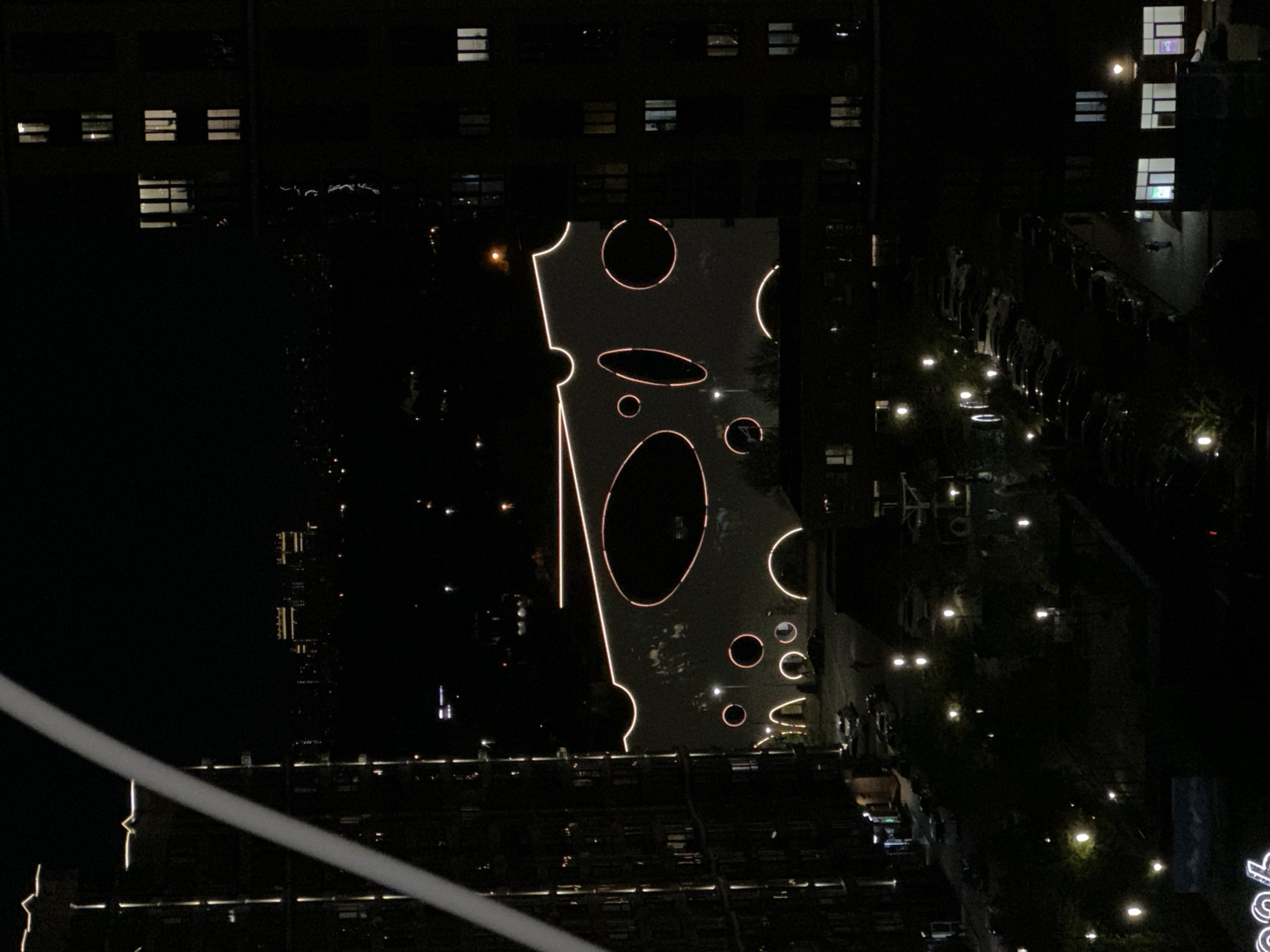 Here's a description of the image:
The photograph, taken at night, showcases an aerial view of a city landscape. The primary subject is a large, uniquely designed structure illuminated with orange lighting. It is positioned between two buildings, resembling a tall slice of cheese with several large circular cutouts. The orange lights highlight the edges of the "cheese" and the perimeters of the holes.
To the right of the structure, a park or garden area is visible. There are trees, pathways, and numerous small lights scattered across the terrain. These lights likely serve as illumination for the park. Rows of parked cars are also visible.
To the left of the cheese slice, a dark, unlit space extends, contrasting with the brightly illuminated structure and the park. Buildings surround the central element, and windows in these buildings are visible, some with lights on.
The image also features a faint white diagonal line in the lower left corner, which appears to be a part of another structure or object, but it is not clearly defined.
Based on the context provided, it's assumed that this scene is located in the city of Krasnodar, Russia.