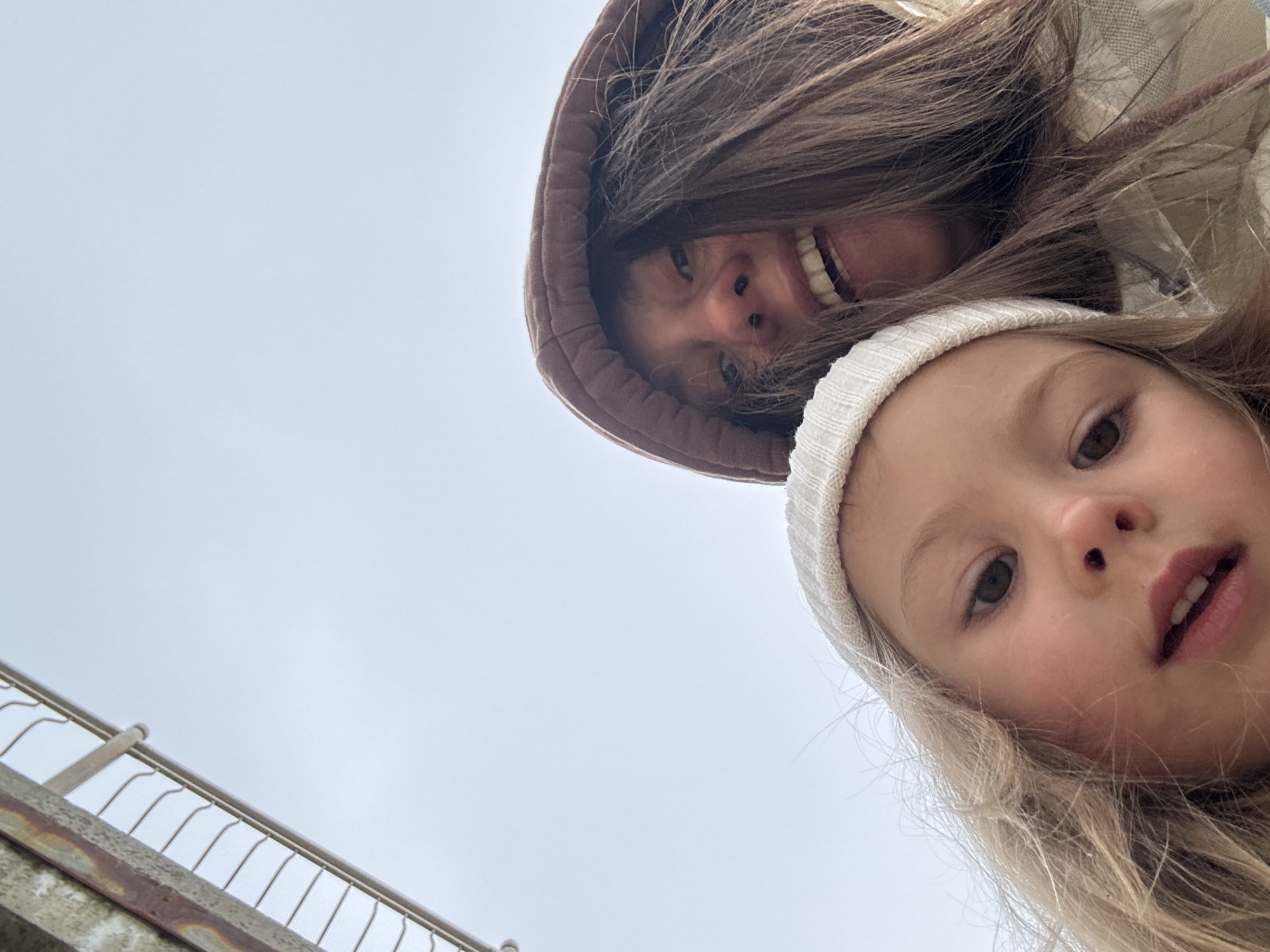 Two individuals are visible in the frame, an adult and a child, angled from below. The adult, wearing a brown hooded jacket, is partially obscured by their hair. The child, wearing a white beanie, is positioned below the adult. The background is dominated by a light gray sky. In the lower-left corner, there's a railing, implying a bridge or elevated walkway. Given the information, the location is likely in the city of Zelenogradsk, Russia.