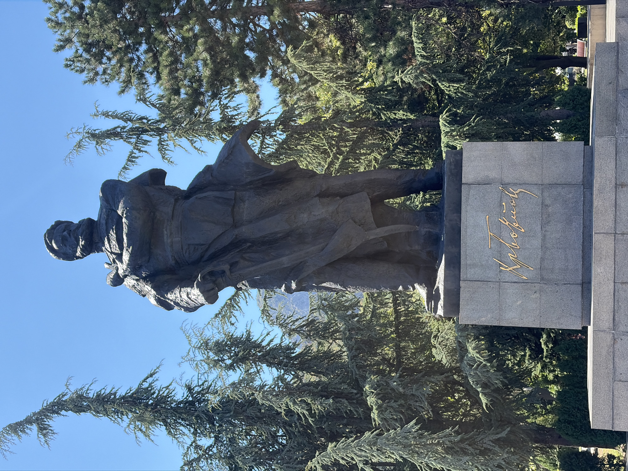 A full-body bronze statue of a man stands on a granite pedestal in Vratsa, Bulgaria. The man is depicted with a beard and is wearing a military-style uniform with a long coat. The inscription on the pedestal is in gold, stylized Cyrillic script. The statue is situated outdoors, with evergreen trees and a clear blue sky forming the background. In the distance, mountains are visible. The scene appears to be a public monument within a park or square.