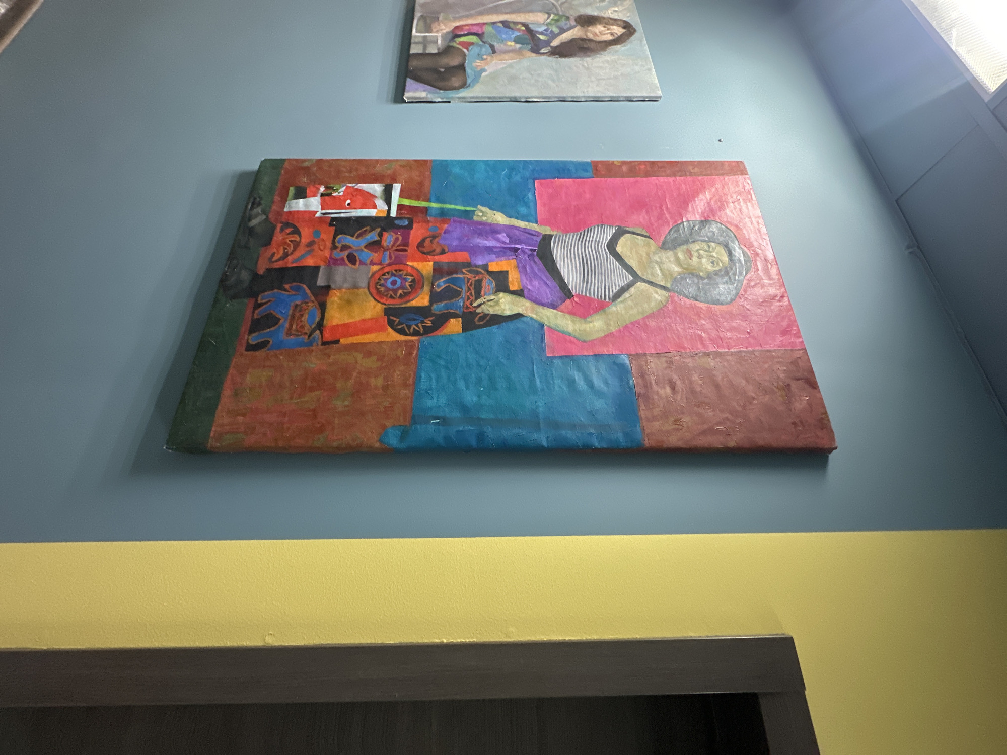 The photograph shows a wall with three distinct sections. The bottom section, colored yellow, is topped by a dark gray horizontal line. Above the yellow section, the wall is painted light blue. Two paintings are mounted on the light blue section of the wall. The lower painting depicts a reclining figure with a hat wearing a striped top. The person is lying diagonally across the painting, which is a mix of geometric shapes and colors including brown, blue, and pink. Above this is a smaller, rectangular painting showing a reclining woman in a dress or outfit with floral or patterned details. The location appears to be an interior space. No evidence suggests this is in Saint Petersburg, Russia.