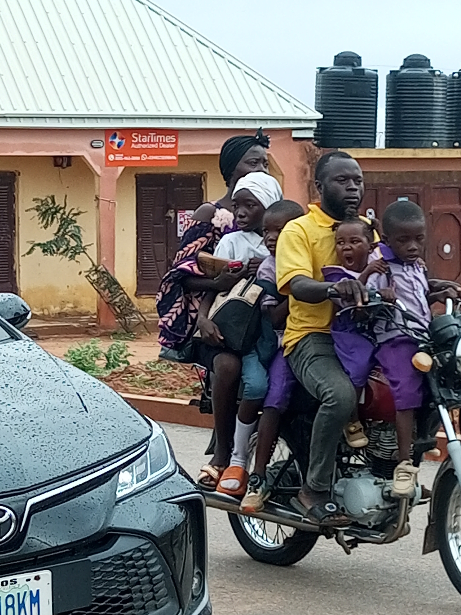 In Gombe, Nigeria, a group of people, including at least one adult and multiple children, are riding on a motorcycle. The adult, a man wearing a yellow shirt, is driving, with children seated in front of him and behind him. One child is seated directly in front of the driver, a young girl with her mouth open, wearing a purple dress. Another child, a boy, is seated behind the driver, wearing a purple shirt and shorts. A woman, partially obscured, is seated behind the driver, holding an infant. The motorcycle is black and red. In the foreground, the front of a dark-colored car is visible. Behind the motorcycle and the people, there is a building with a light-colored roof and orange accents. Two large black water tanks are visible on top of the building. The environment appears to be a street or road.