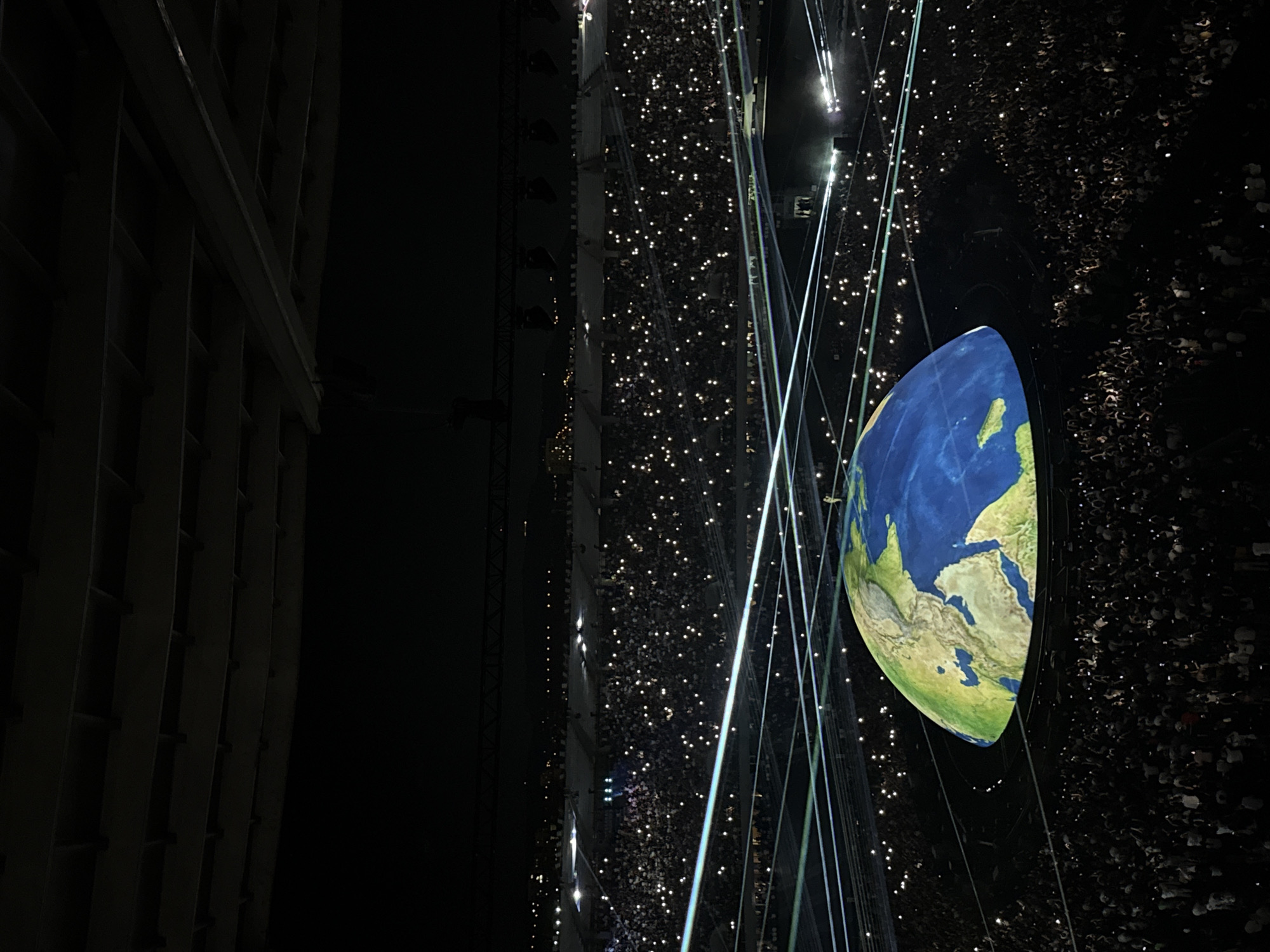 The image shows a large stadium filled with a crowd of people holding up their phones with lights illuminated. A massive, semi-spherical projection of the Earth is displayed in the center of the stadium, with a green and blue representation of continents and oceans. Several illuminated lines, possibly laser beams or light projections, are visible crisscrossing the stadium above the audience and the Earth projection. The stadium structure is visible in the upper portion of the image, with rows of seating occupied by the audience. The overall scene suggests a large-scale outdoor event, likely a concert or a ceremony, at night. The location is identified as the Boris Paichadze Dinamo Arena in Tbilisi, Georgia.