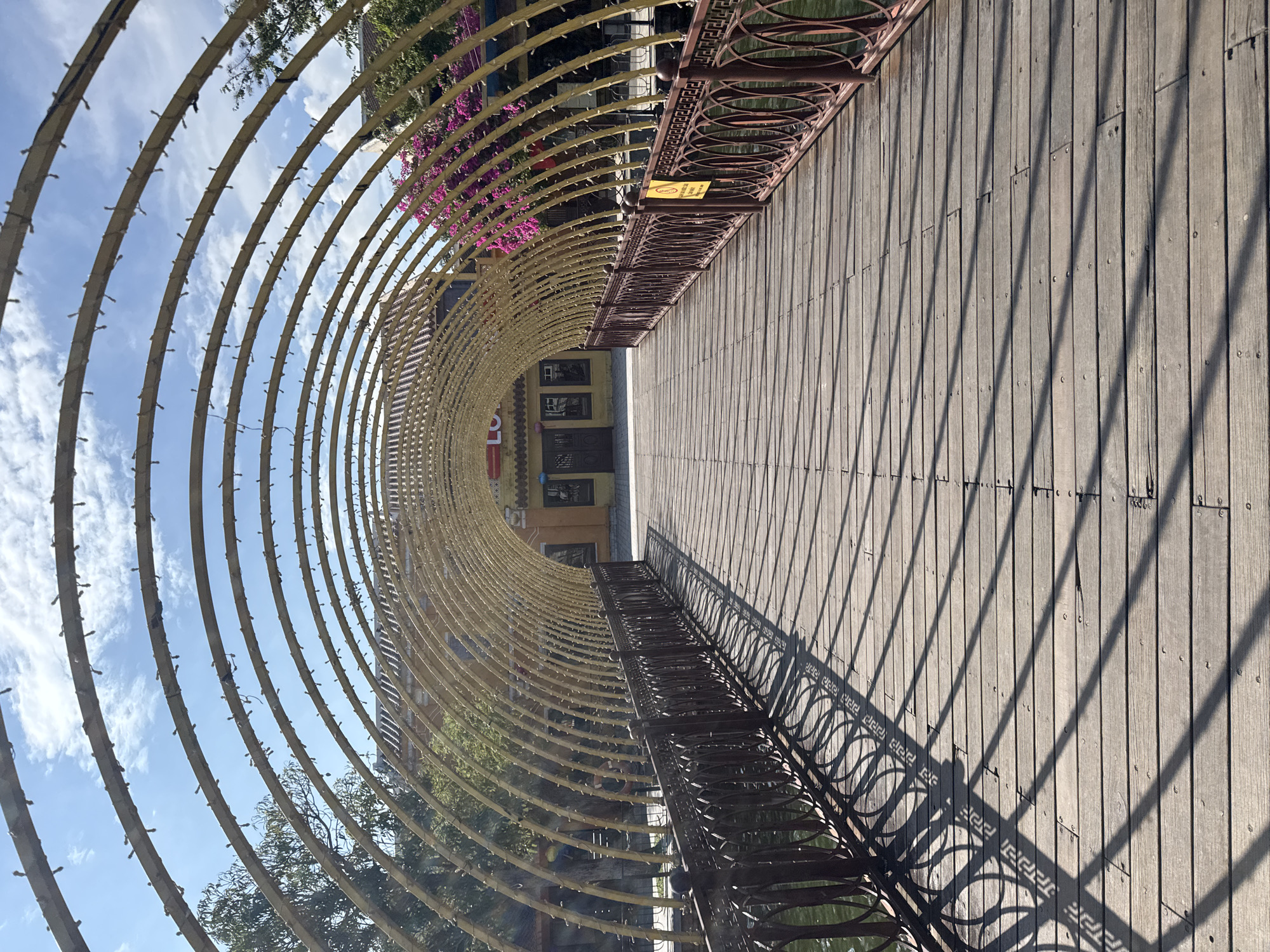 The image depicts a pedestrian bridge in Đà Nẵng, Vietnam. The bridge has a wooden deck with a dark metal railing along the sides. Arched, lit structures curve above the bridge, creating a tunnel effect. Shadows from the railings and arches cast linear patterns on the wooden surface. In the background, there is a building with multiple windows and a visible storefront. Parts of trees with green foliage are visible alongside the bridge. The sky is partially visible through the arches, showing a blue sky with scattered white clouds.