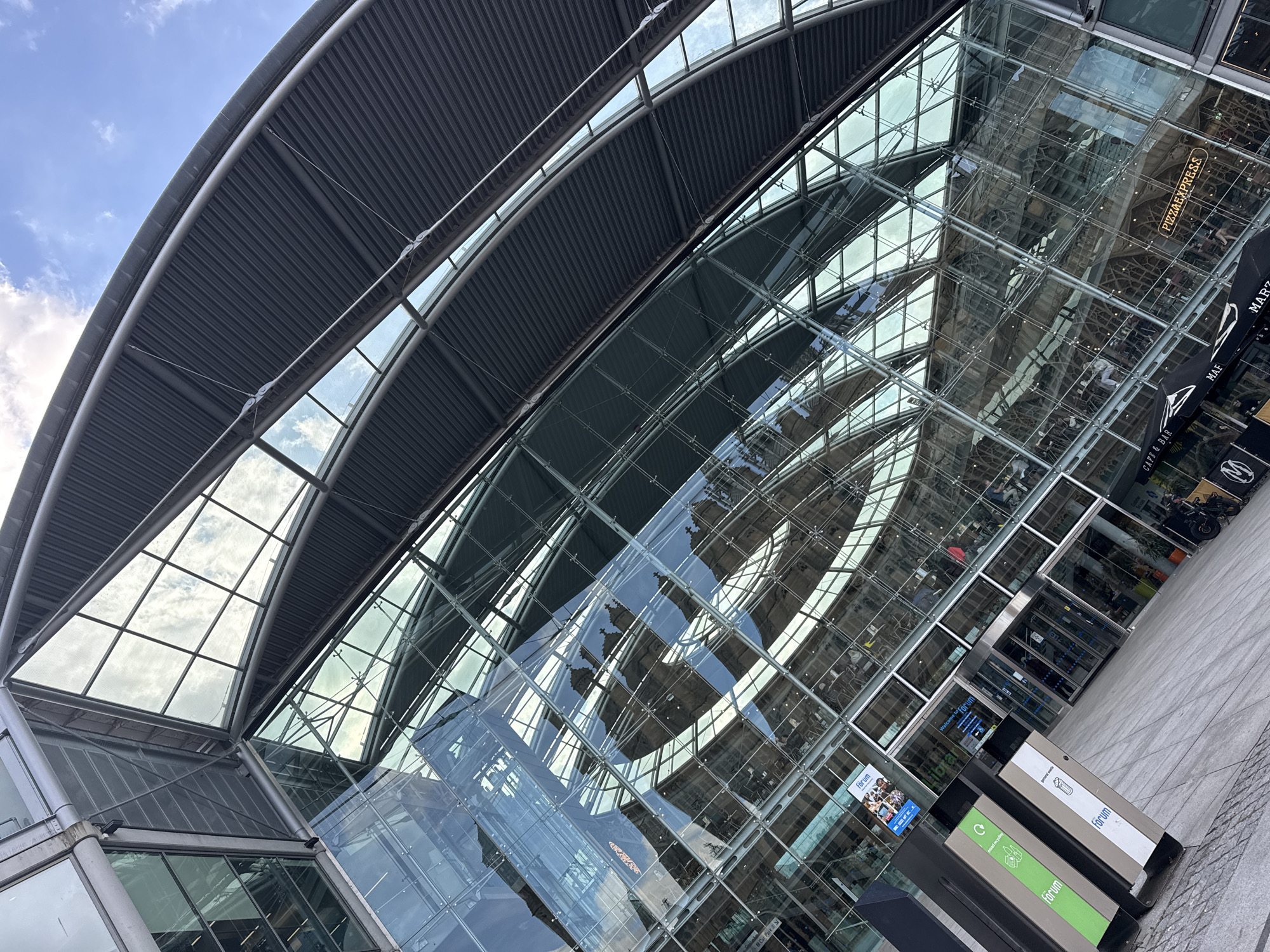The image depicts the Forum building in Norwich, UK, viewed from an upward angle. The building has a curved gray metal roof structure with exposed supports. The facade is largely glass, reflecting the sky and the surrounding buildings. A Pizza Express sign is visible in one of the reflected scenes. At the base of the building, there are public waste bins labeled "Forum." The overall architectural style is modern and geometric. The sky is pale blue with scattered clouds.