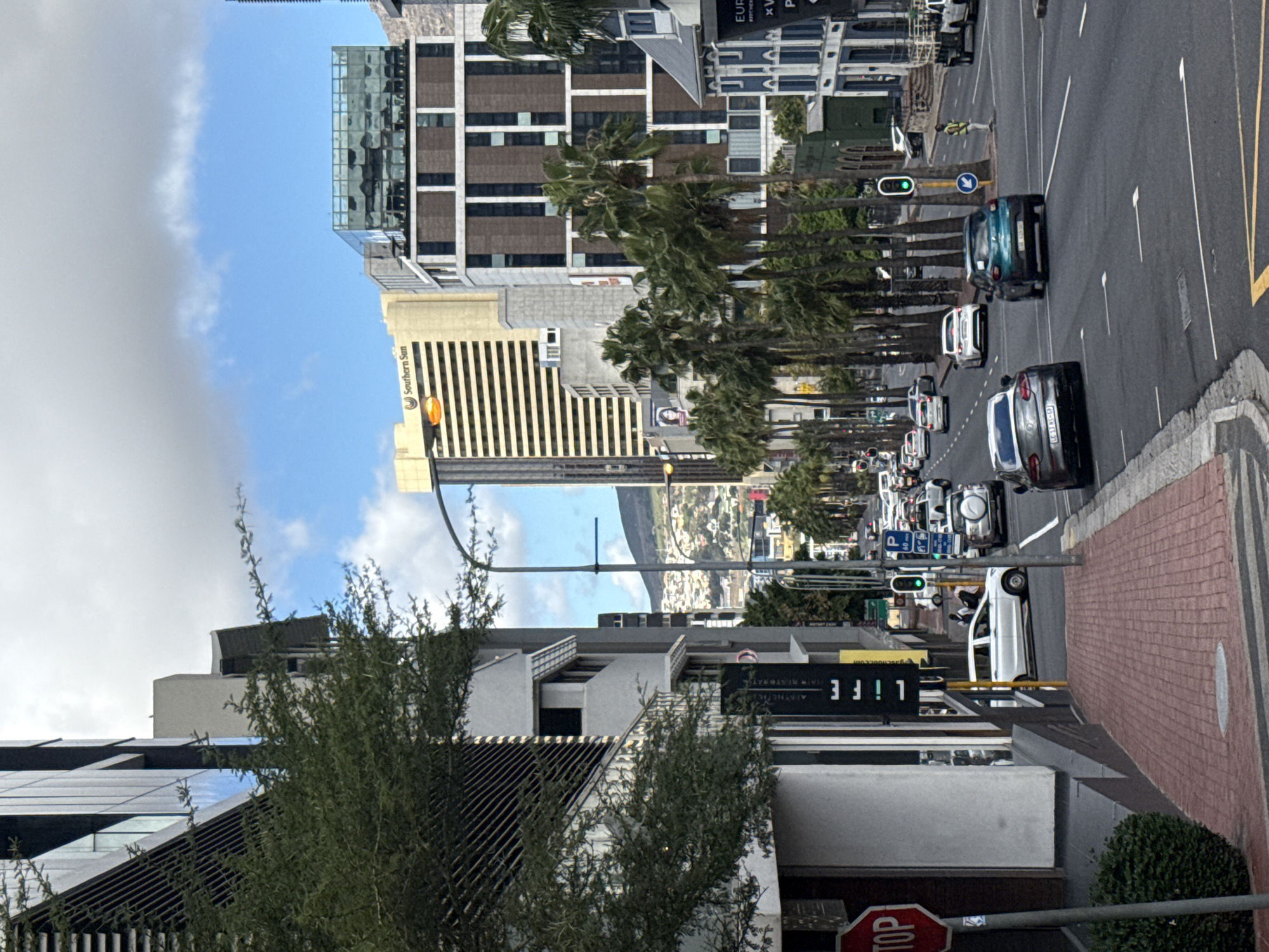 This is a street view in Cape Town, South Africa. The photo shows a road with several cars moving in the same direction. Palm trees line both sides of the road. Buildings of various heights flank the street, including one identified as "Southern Sun". In the foreground, there is a stop sign and a sign reading "LiFE", indicating a business or establishment. The sky above is partly cloudy, and in the distance, there is a view of a mountain or hill. The overall environment is urban and appears to be a commercial district.