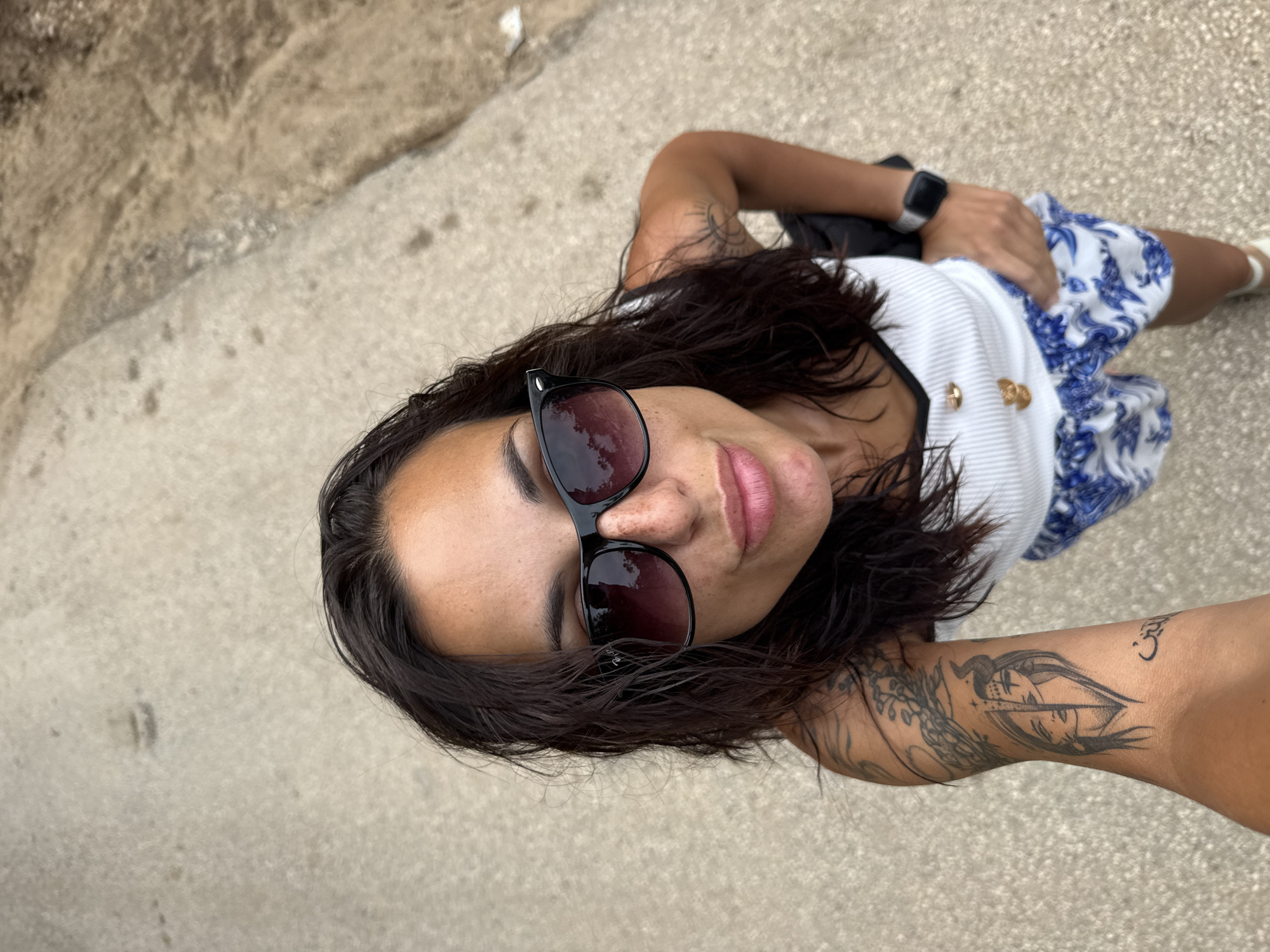 A woman with long, dark, wet hair and sunglasses takes a selfie from a high angle. She is wearing a white ribbed tank top with gold buttons and blue and white patterned shorts. She has tattoos on her left arm, including a design of two faces and a sun. A smartwatch is visible on her left wrist. She is standing on a paved surface, likely a street or pathway, with a sandy or gravelly area to her right. The overall lighting suggests an outdoor setting during the daytime. There is no identifiable information to suggest the location is in the city of Pleven, Bulgaria.