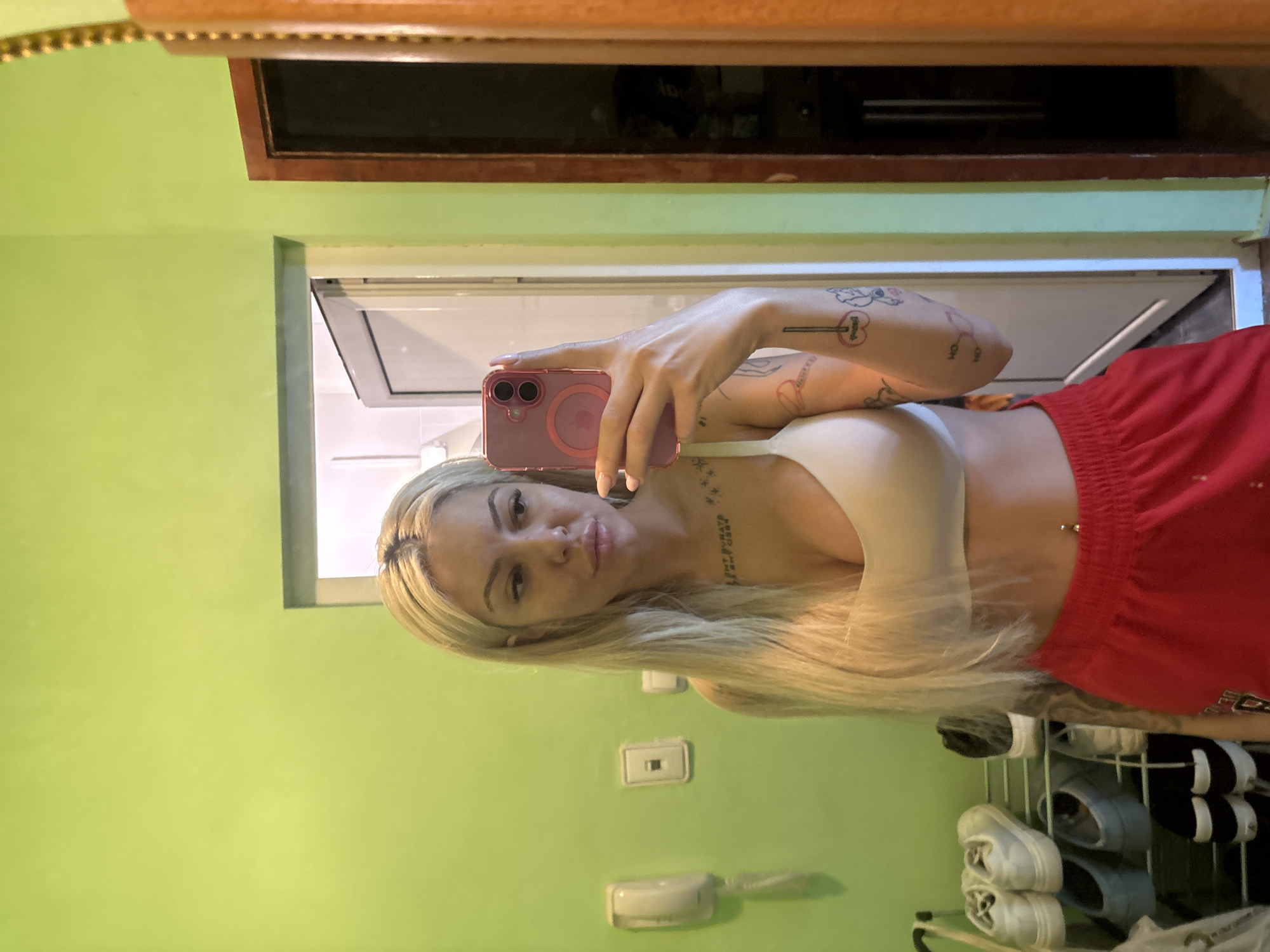 A blonde-haired woman is taking a selfie in a room with green walls. She is wearing a white bra and red shorts. There is a mirror above her with a wooden frame. Behind her, there is a doorway leading to another room that appears to have a white interior. A shoe rack with multiple pairs of shoes is visible in the lower right corner of the image. There are electrical outlets and a wall-mounted telephone on the wall. There is no contextual information suggesting the image was taken in Plovdiv, Bulgaria.