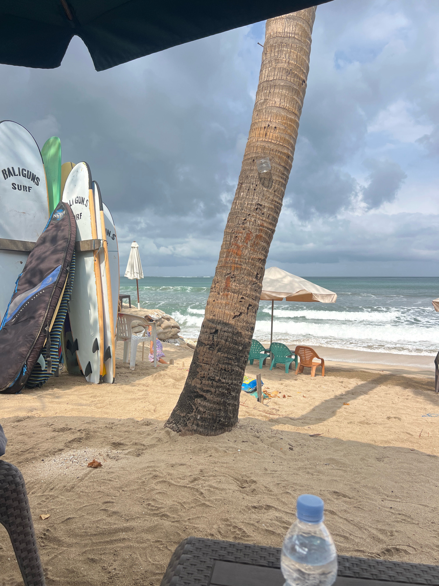 This is a photograph of a beach scene in Kuta, Indonesia. In the foreground, a water bottle with a blue cap rests on a dark, woven surface, likely a table. The sand is a light tan color and appears undisturbed in some areas, with footprints and ripples in others. To the left, a portion of a dark, open umbrella is visible, casting a shadow. Several surfboards are propped up against a wooden structure or wall on the left side of the image, some with brand names like "BALIGUNS SURF" visible. A large palm tree trunk dominates the center-right of the frame, its rough bark textured and weathered. Behind the palm tree, a beach umbrella shades a few plastic chairs – two green and one orange. The ocean is visible in the background, with moderate waves breaking on the shore under a cloudy sky. The water is a greenish-blue hue. There is a white umbrella near some chairs and a table set back from the water.