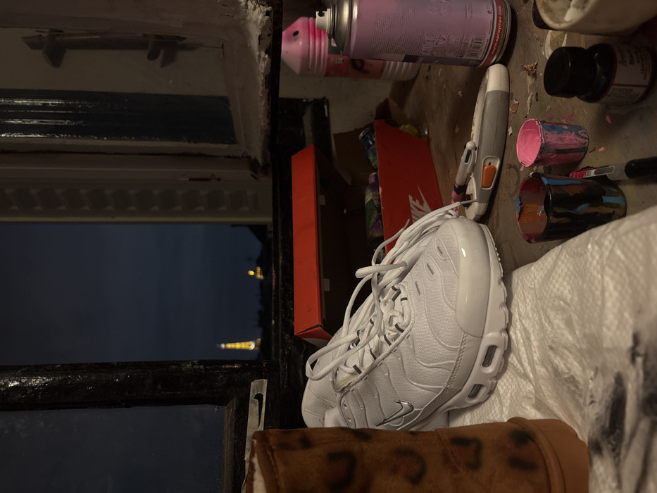 The image depicts an indoor workspace at dusk or night, featuring a window that frames the illuminated Eiffel Tower in the distance, identifying the location as Paris, France.
In the immediate foreground, a white Nike Air Max Plus (TN) sneaker with untied laces rests centrally on a white cloth or paper towel. To its left, the upper portion of a brown boot with dark, irregular markings is visible. Behind the sneaker, an open red box, likely a Nike shoe box, contains various items.
To the right of the sneaker, a collection of art and craft supplies is arranged on the surface. These include a grey utility knife with an orange slider, a multi-colored pen, two small, paint-splattered containers (one predominantly black and blue, the other pink and blue), a black marker, a small black bottle containing a red liquid, and two spray paint cans—one pink and one lavender, with the lavender can labeled "spraymax" and showing a capacity of 400ml.
The window frame is dark and appears weathered. The sky beyond is a deep blue, with the prominent, brightly lit Eiffel Tower positioned off-center to the left, and a fainter distant light visible to its right.