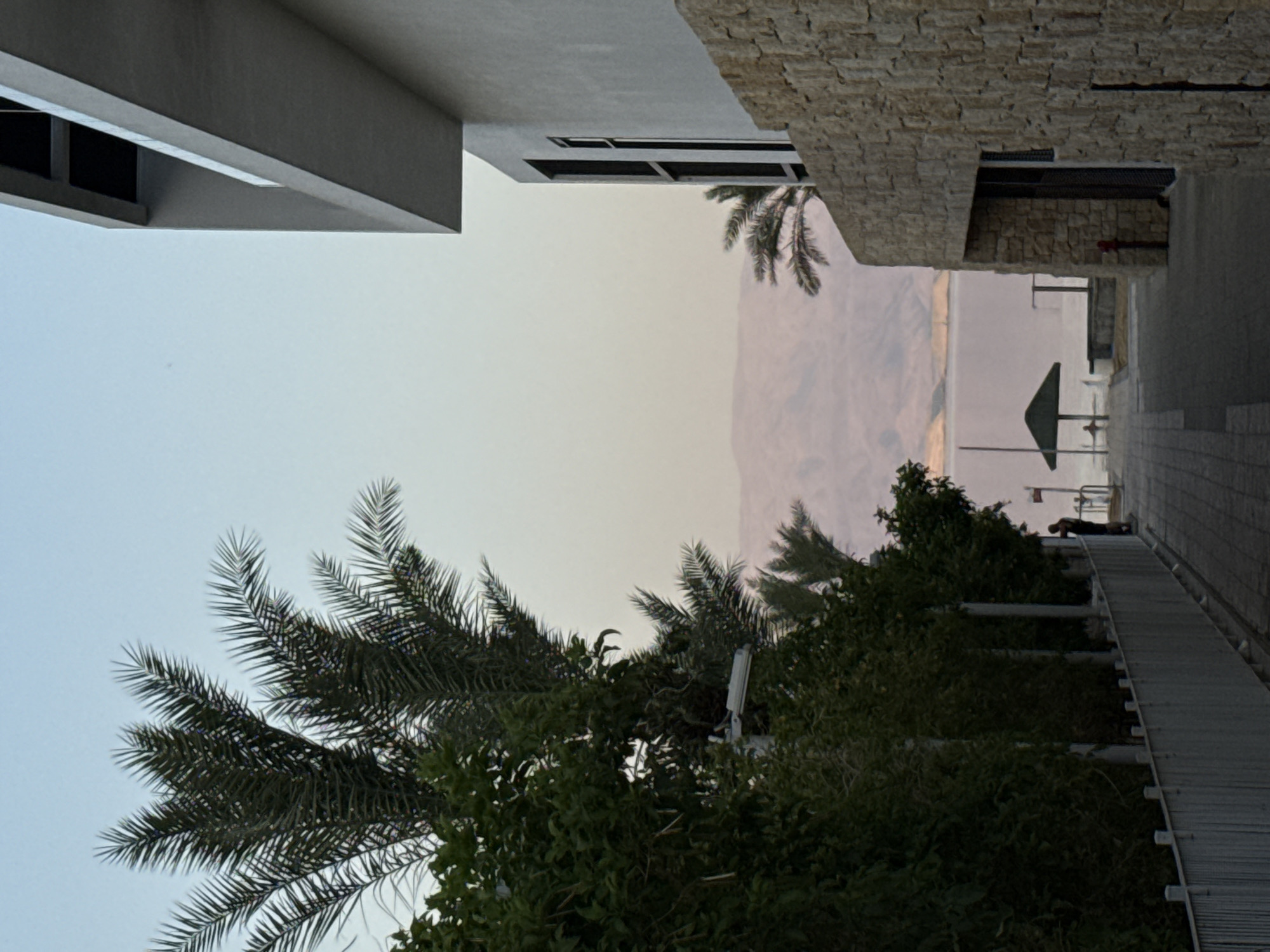 A serene view of the Dead Sea at dusk, framed by lush palm trees and modern architecture, is captured in this image. The sky is a soft gradient of blue and pink, casting a warm glow over the scene. The Dead Sea stretches out in the background, its calm waters reflecting the muted colors of the sky. In the distance, the mountains of Jordan are visible, their contours softened by the twilight.
In the foreground, a pathway lined with palm trees leads to the water's edge. A solitary figure stands on the pathway, looking out at the sea. A green umbrella, providing shade from the sun, is set up on the beach. To the right, a contemporary building with a stone facade stands tall, its clean lines contrasting with the natural beauty of the landscape. The building's windows offer glimpses of the interior, hinting at a place of rest and relaxation.
The overall atmosphere is one of tranquility and peace. The image evokes a sense of calm and invites the viewer to immerse themselves in the serene beauty of the Dead Sea. This scene is likely located in Tamar Regional Council, Israel, a region known for its stunning natural landscapes and proximity to the Dead Sea.