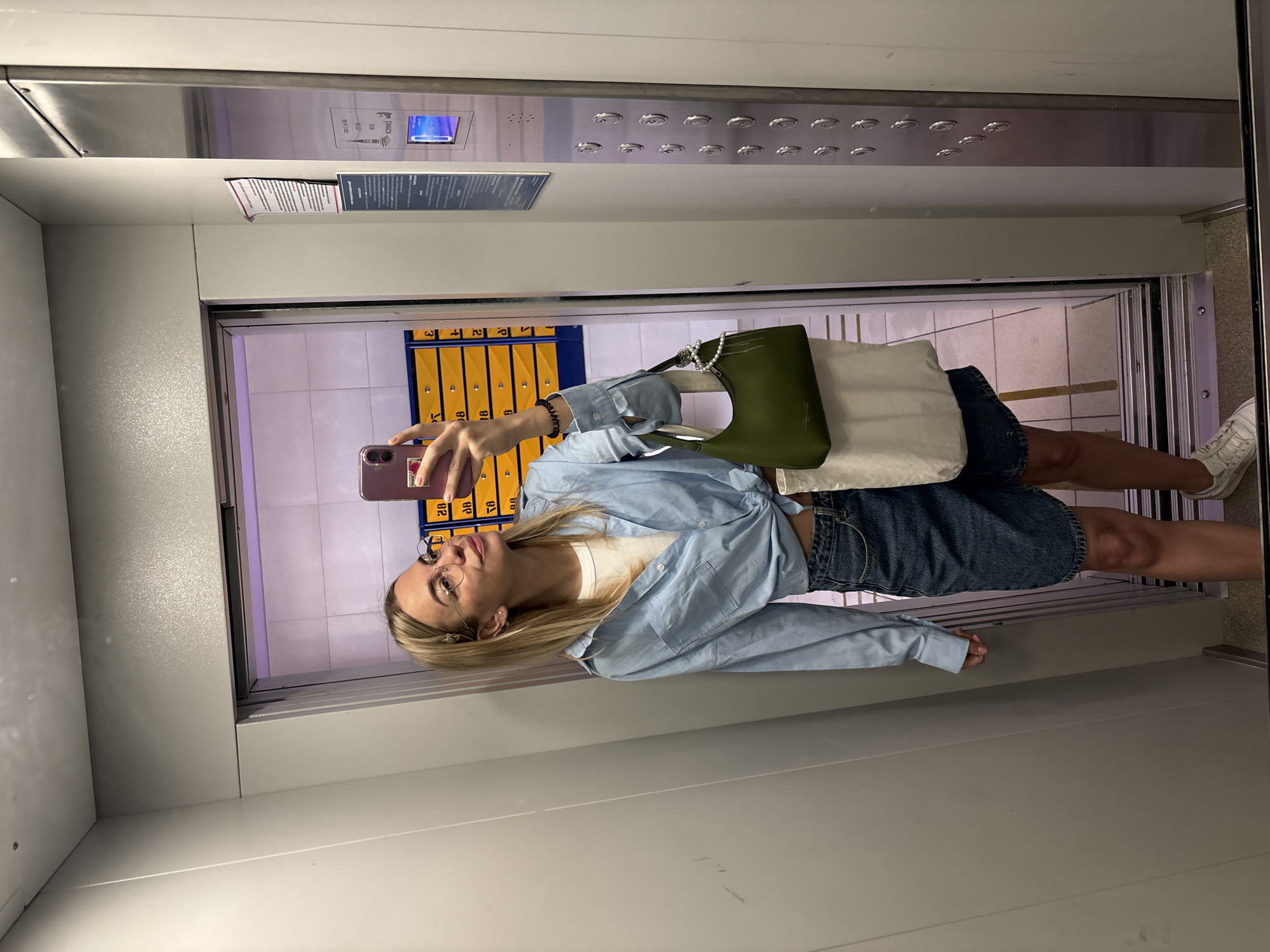 A woman takes a selfie in a mirrored elevator. She wears glasses, a white t-shirt, a light blue shirt tied at the waist, and a denim skirt. She holds a pink phone in her left hand. A green handbag and a white tote bag are slung over her shoulder. In the background, behind the elevator door, are rows of yellow lockers. The reflection reveals the elevator's interior, including the control panel. A sign near the top is partially visible. The scene suggests an urban setting, possibly in Krasnoyarsk, Russia.