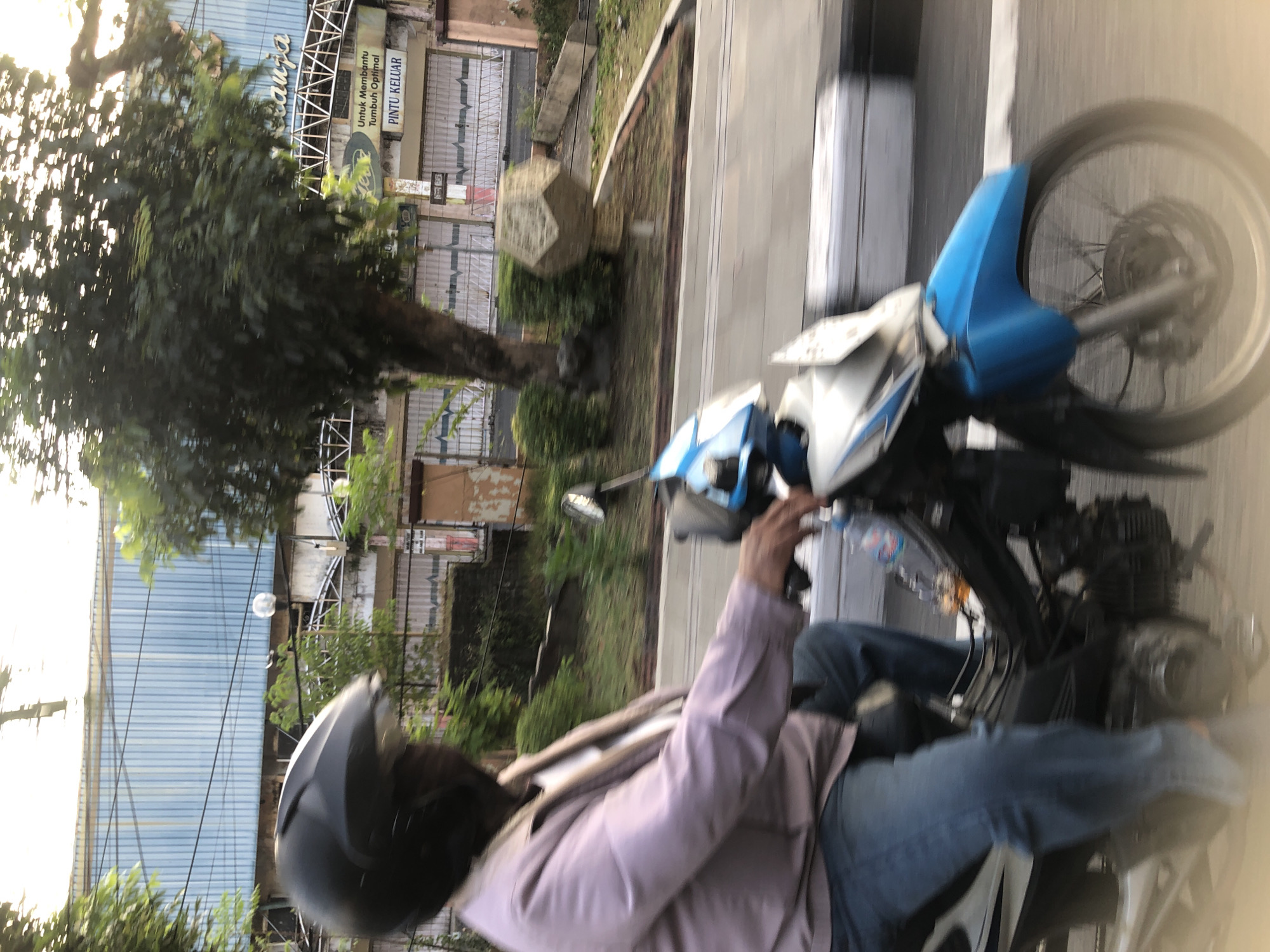 A person wearing a black helmet and a light purple jacket rides a blue and white motorcycle down a street in Larangan, Indonesia. The person is wearing blue jeans and is blurred due to motion. The motorcycle is also blurred, with its front wheel visible. In the background, there is a building with a blue, corrugated metal facade and a white fence. A tree with green leaves is in front of the building. A sign on the building reads "Untuk Membantu Tumbuh Optimal PINTU KELUAR". There is a small, polygonal concrete sculpture in the middle ground, and a concrete barrier with black and white stripes lines the side of the road. The lighting suggests it is daytime.